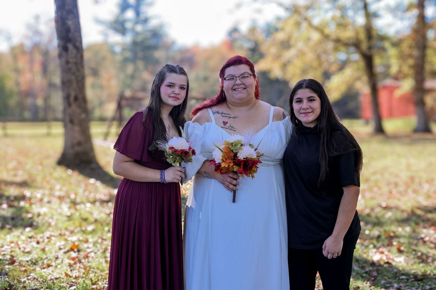Three people stand together outdoors in autumn with colorful fall foliage in the background.