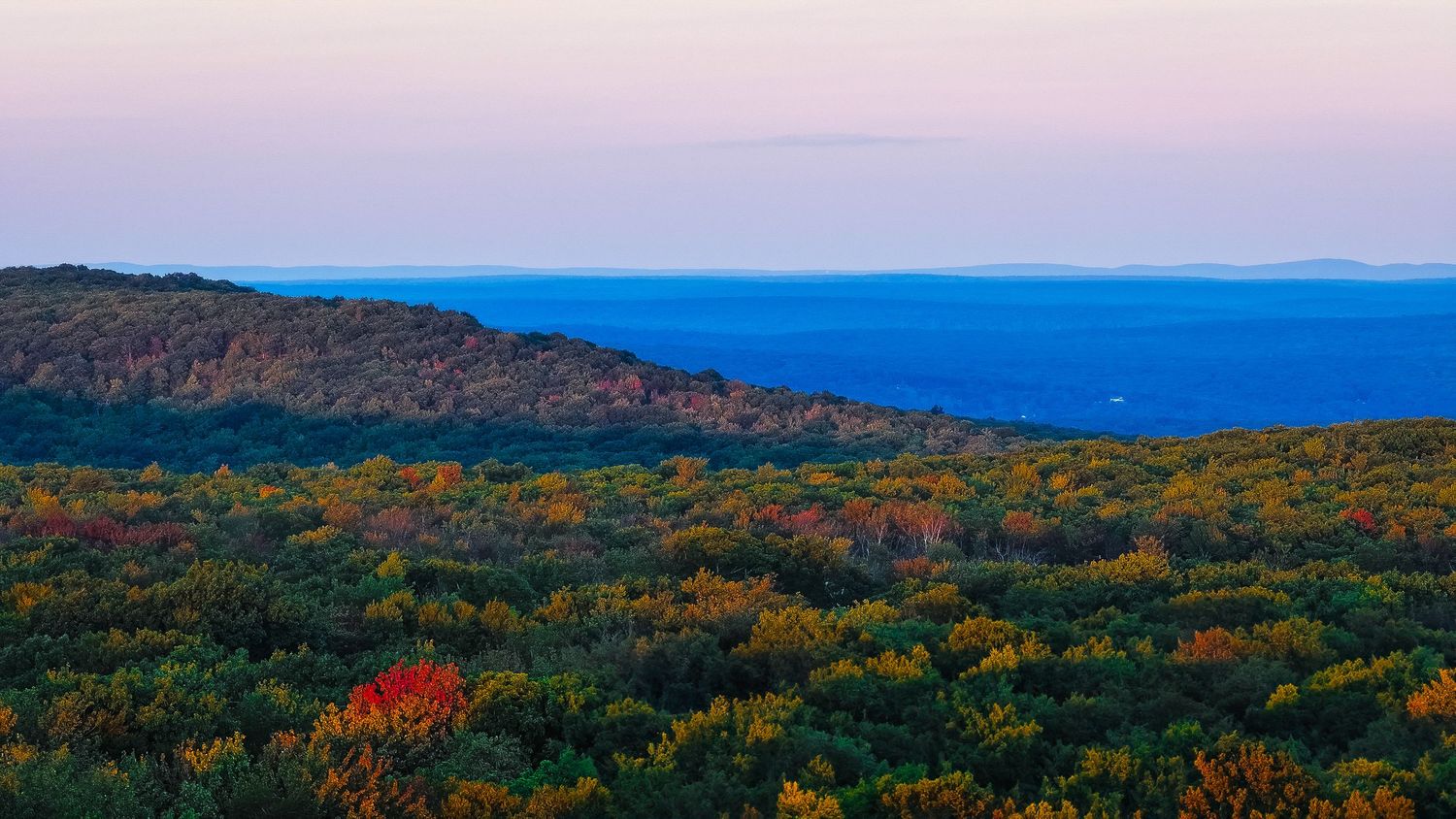A panoramic view of rolling hills covered in vibrant autumn foliage stretches across the horizon at sunset.