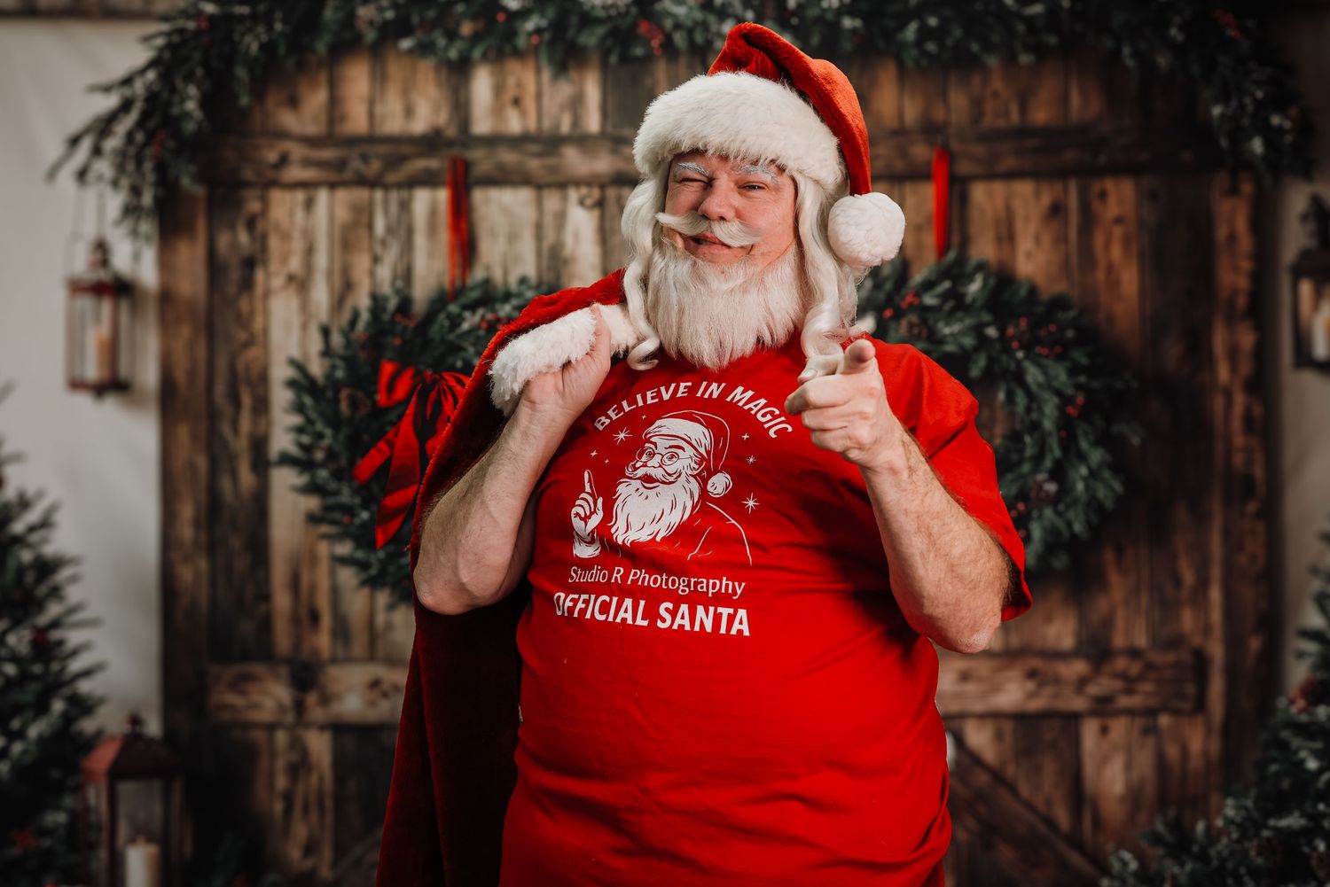 A person wearing a red Santa shirt and hat poses in front of a rustic wooden backdrop for holiday photos.