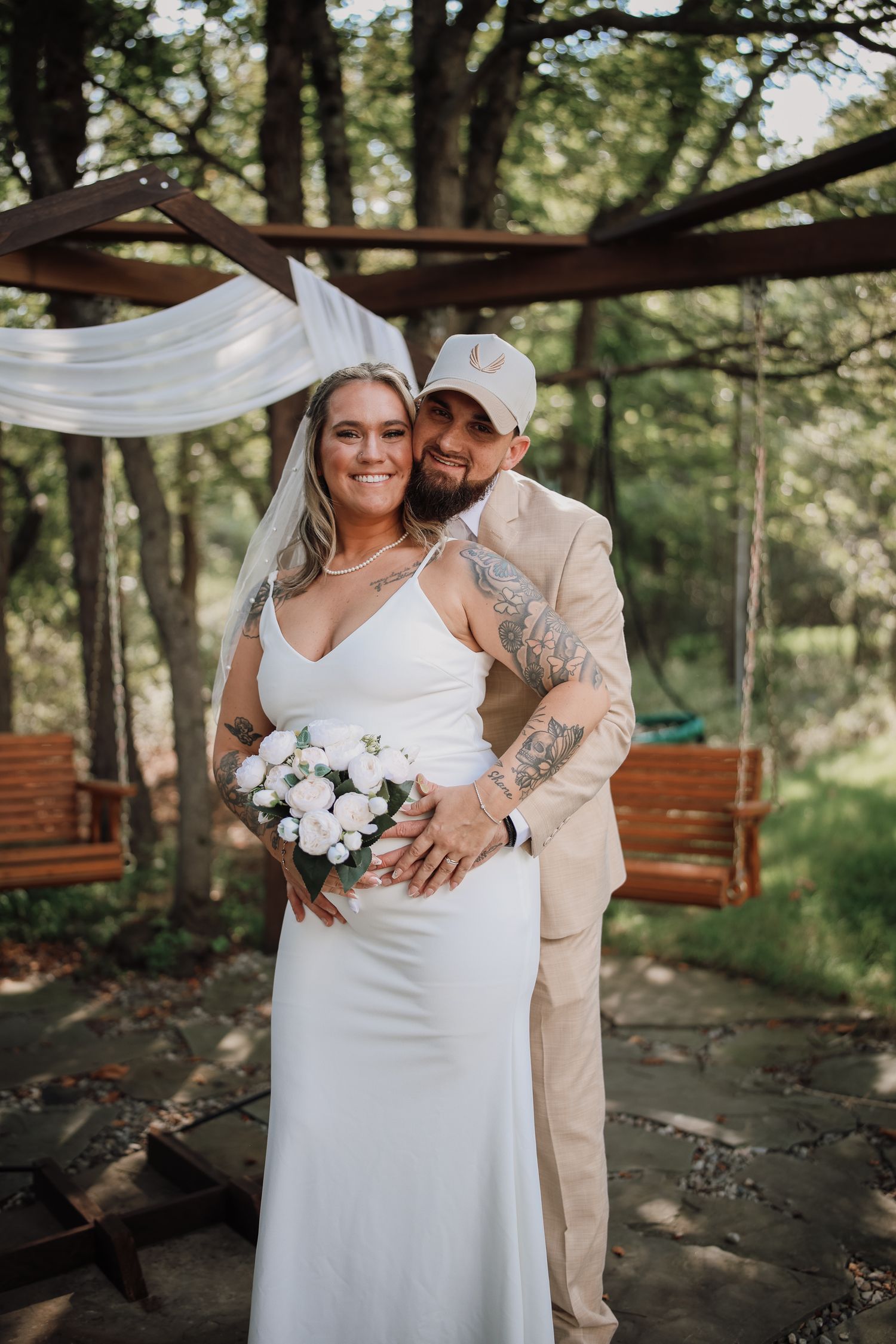 A couple embraces under a wooden pergola during an outdoor wedding ceremony with hanging white fabric and swings.