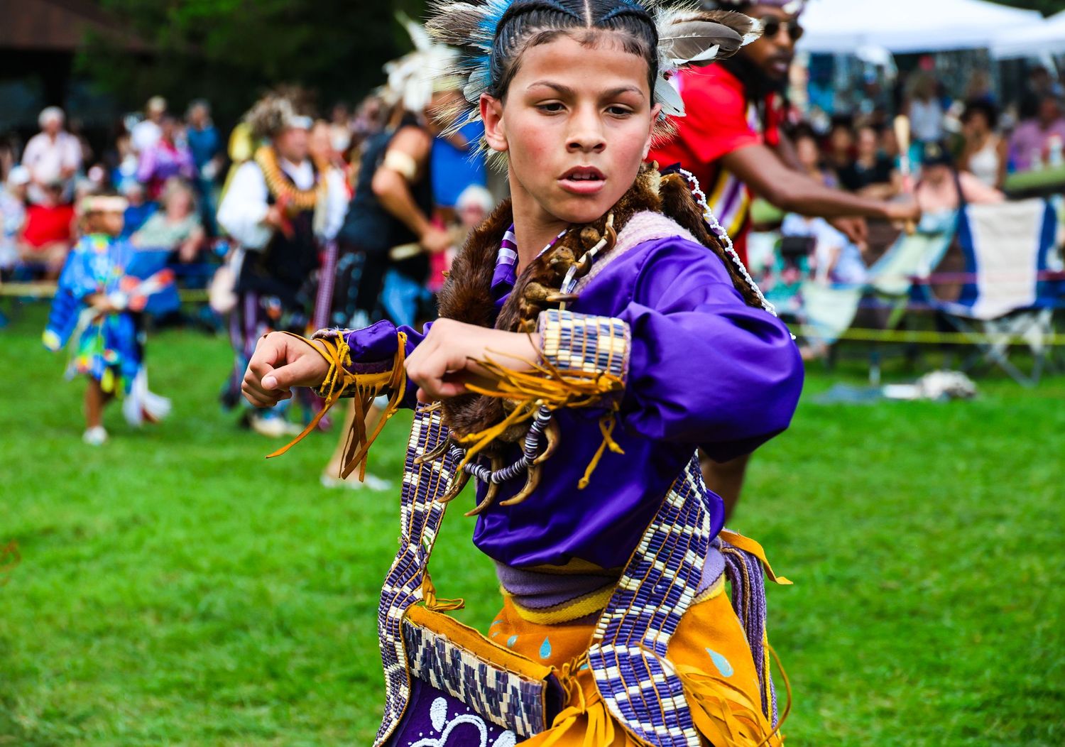 Native American dancers in traditional regalia perform at an outdoor powwow celebration on a grassy field.