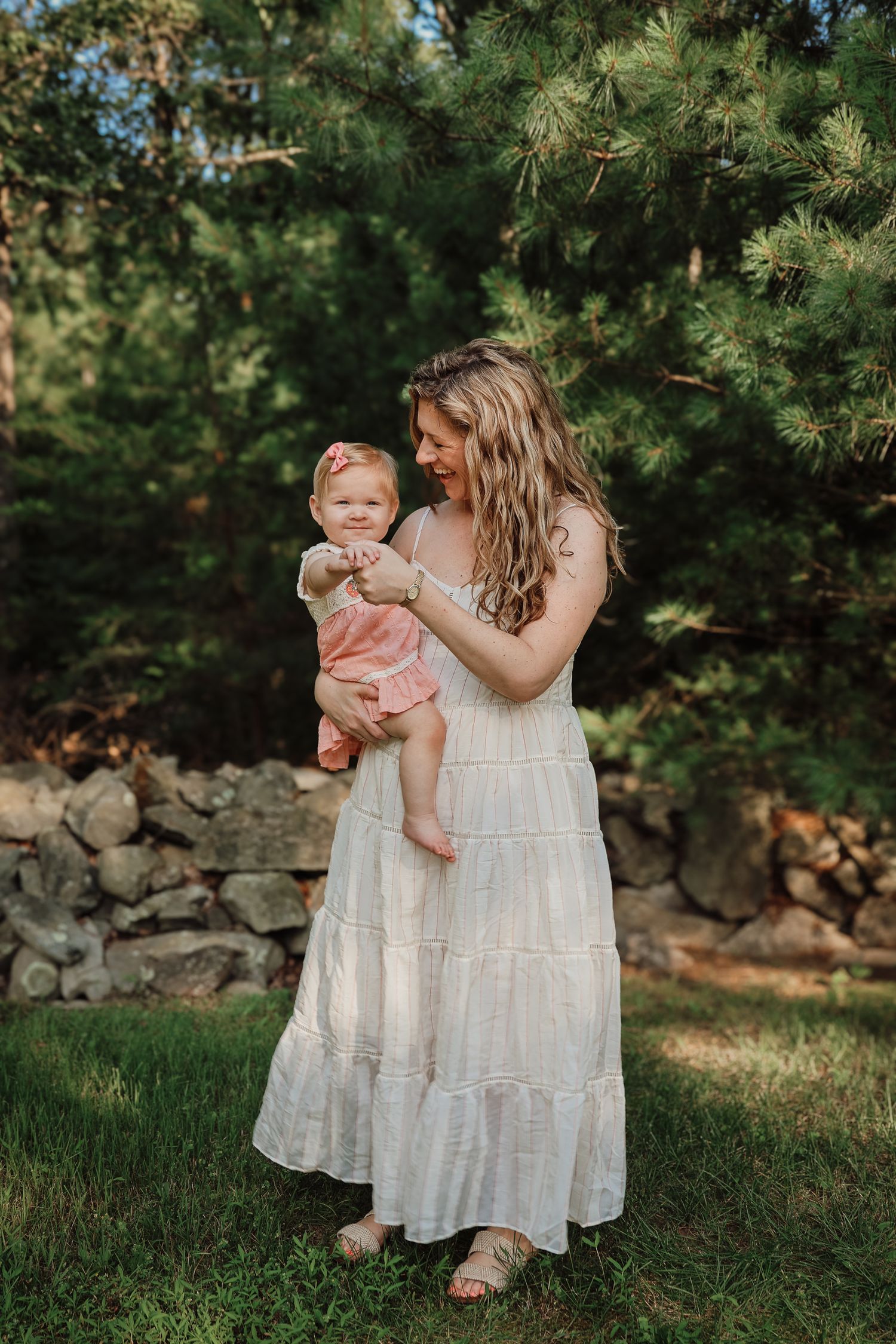 A tender outdoor moment between parent and child in white dresses against a backdrop of evergreen trees and stone wall.