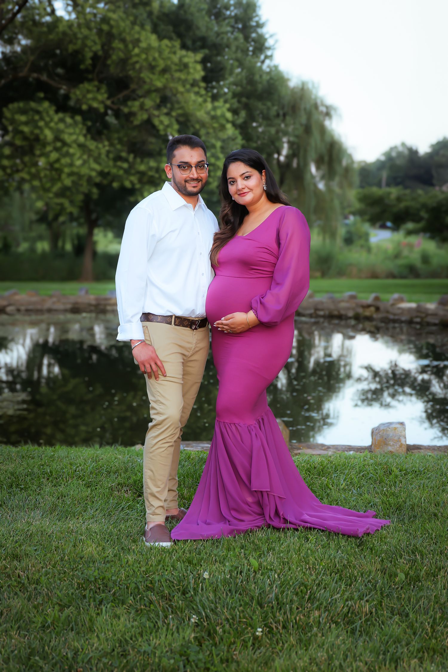 A couple poses for maternity photos by a serene pond on a grassy lawn in a scenic outdoor setting.