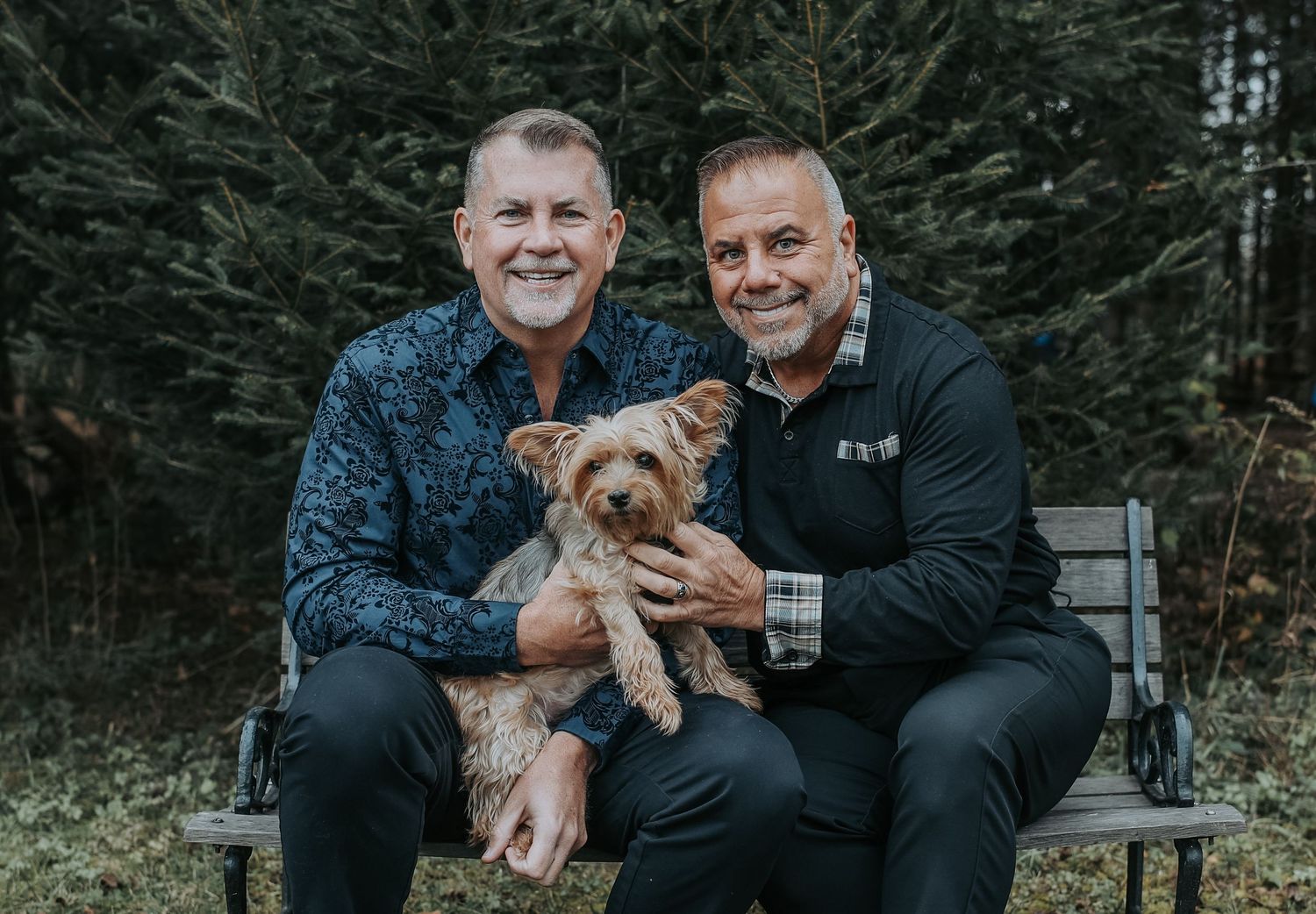 Two people sitting on a bench outdoors with a small brown dog while surrounded by evergreen trees.