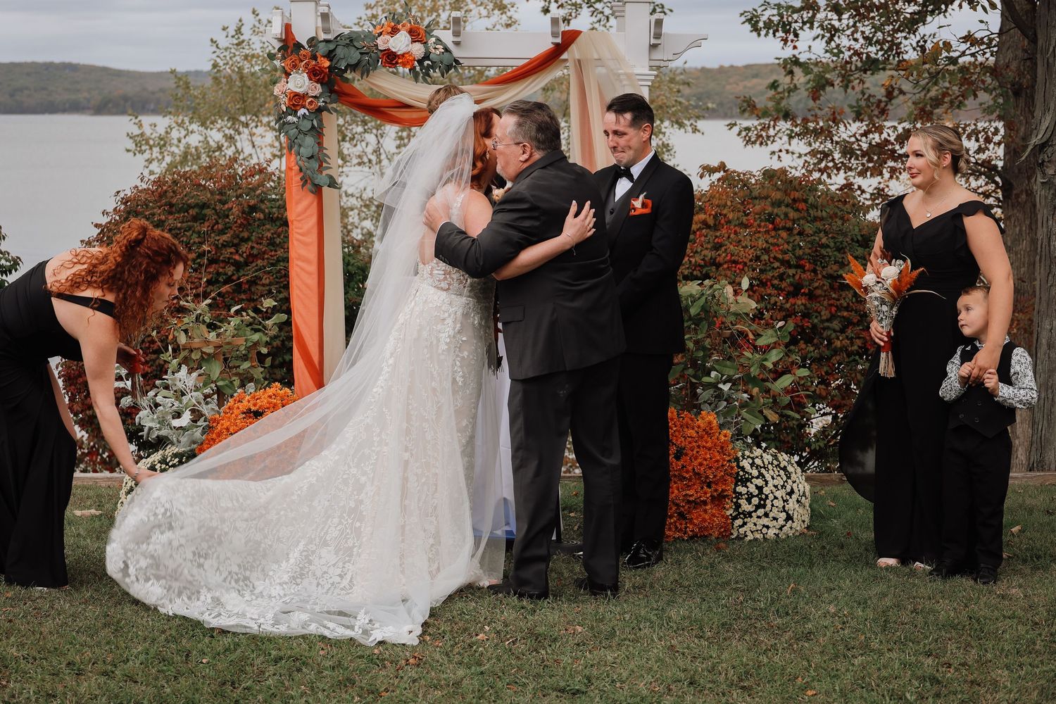 Outdoor wedding ceremony under orange and white arch overlooking lake with wedding party in black attire.
