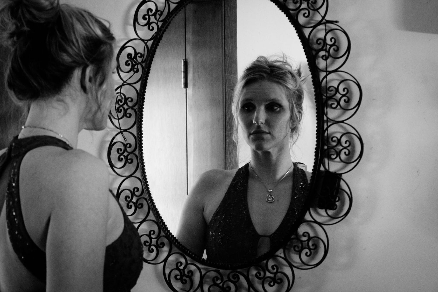 Black and white photo of someone looking into an ornate wrought iron framed mirror against a white wall.