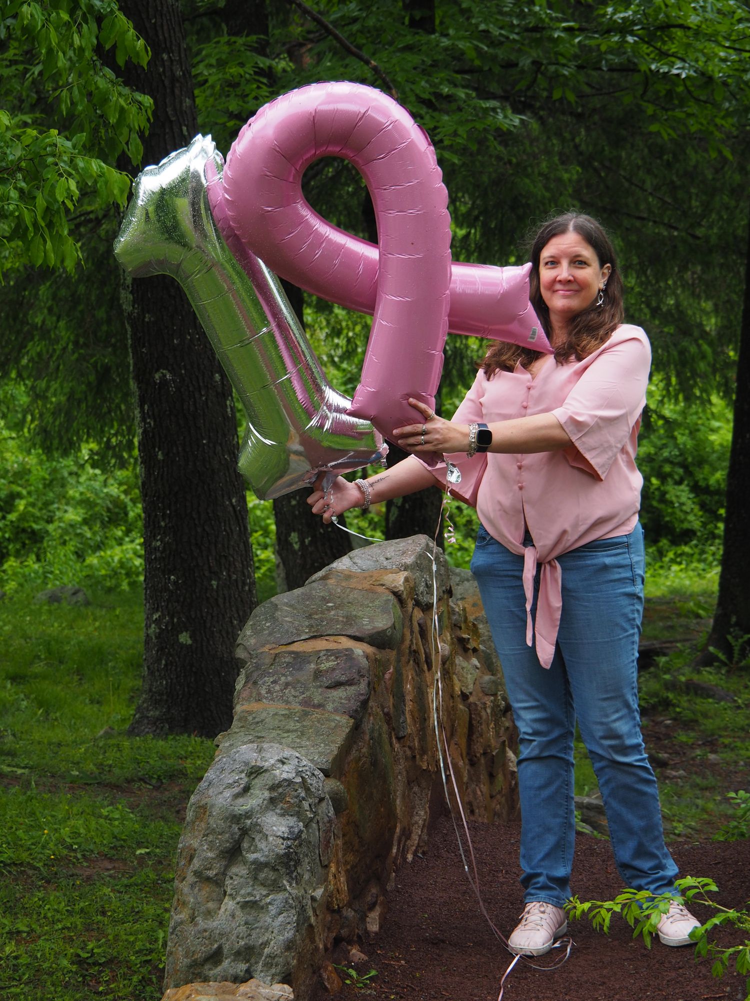 Someone holds a large pink number 2 balloon while standing on rocks in a wooded outdoor setting.