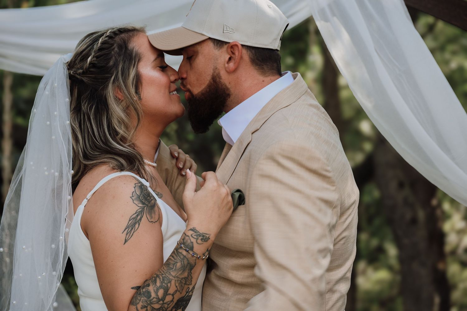 A romantic wedding kiss captured through sheer white curtains in an outdoor garden setting.