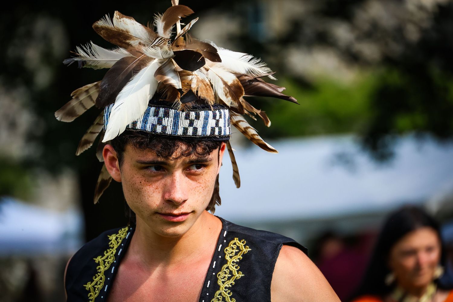 A performer wearing a feathered headdress and traditional regalia participates in a cultural festival outdoors.