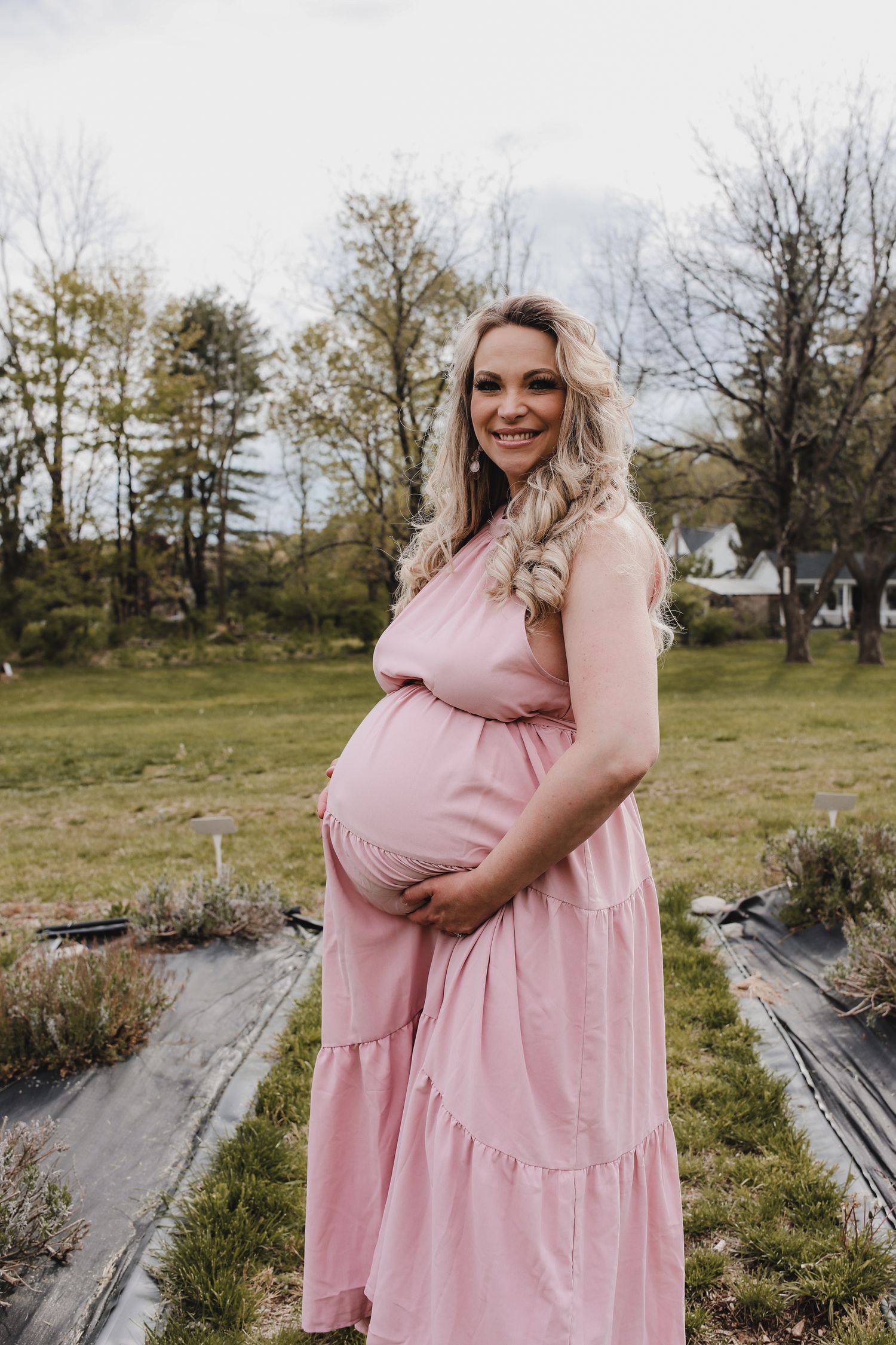 A pregnant woman in a flowing pink dress stands in a grassy field on a spring day.