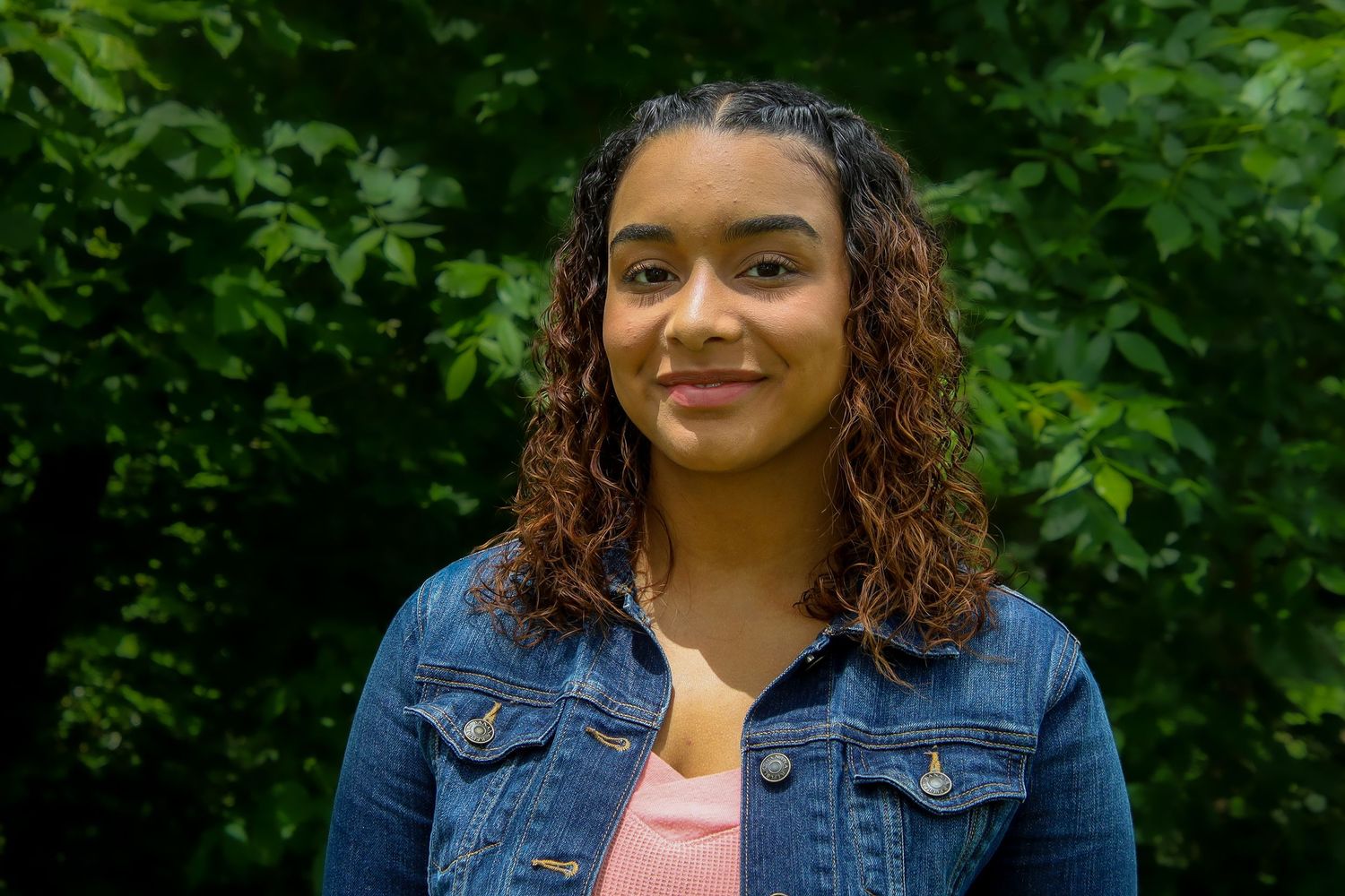 A woman with curly hair wearing a denim jacket and pink top smiles while posing against a green leafy background.
