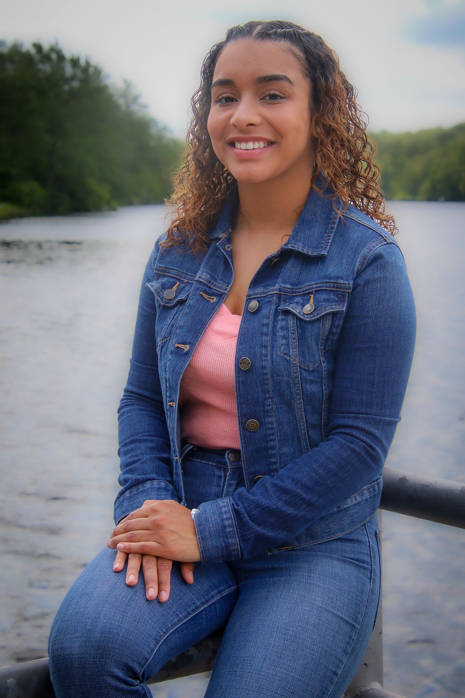 A woman in a denim jacket and coral top sits by a scenic lake on a sunny day.