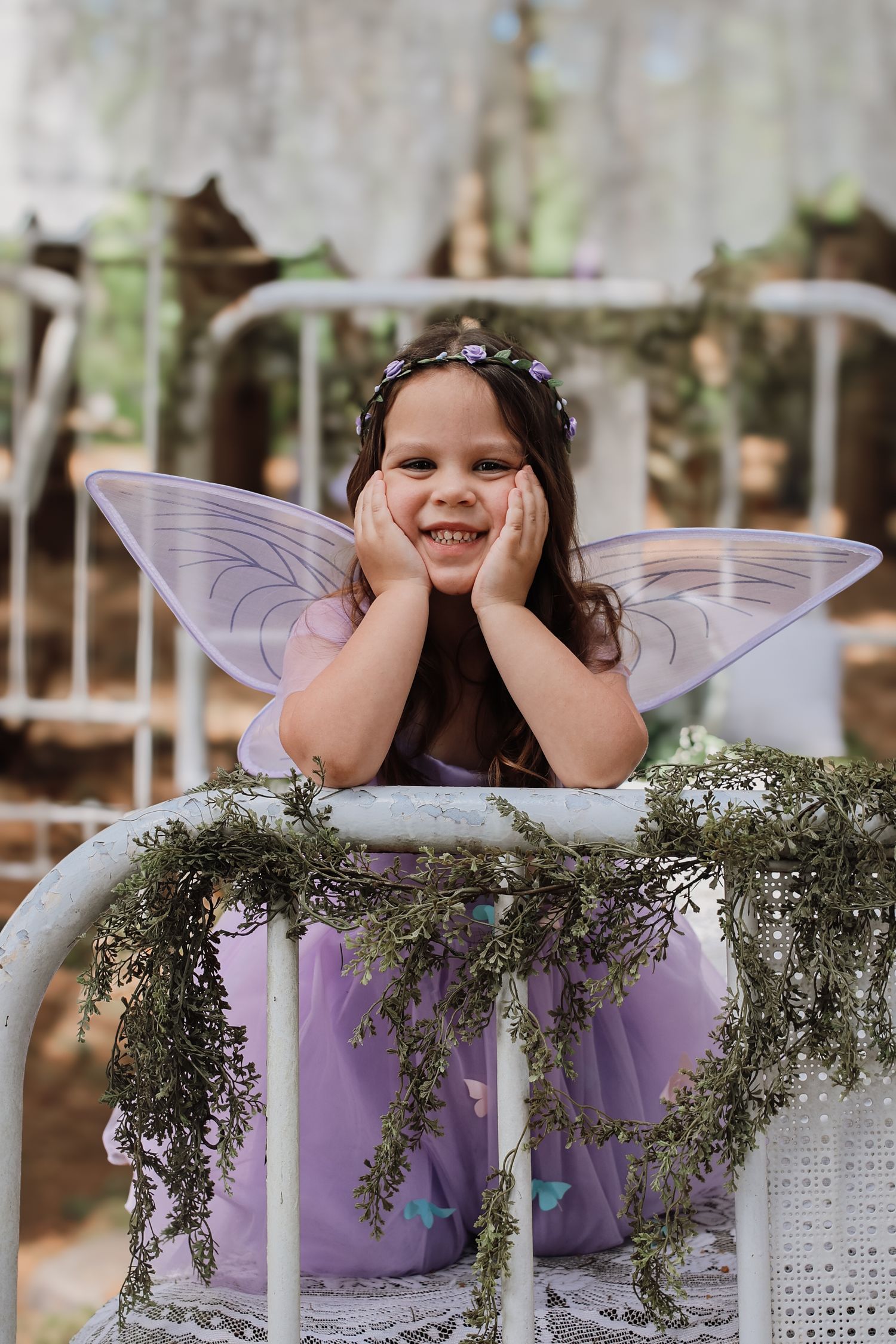 A whimsical fairy-themed photo series showing a child in purple wings resting on white wicker furniture with greenery.