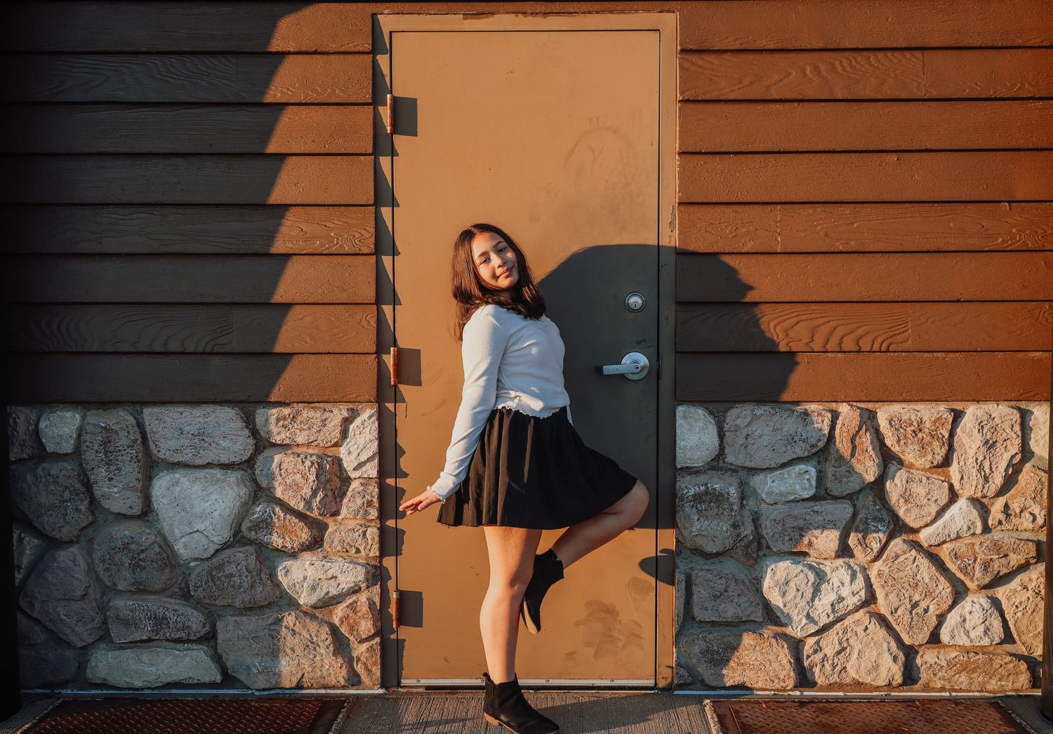 A person in black clothing poses playfully against a rustic wood and stone wall with dramatic shadows.