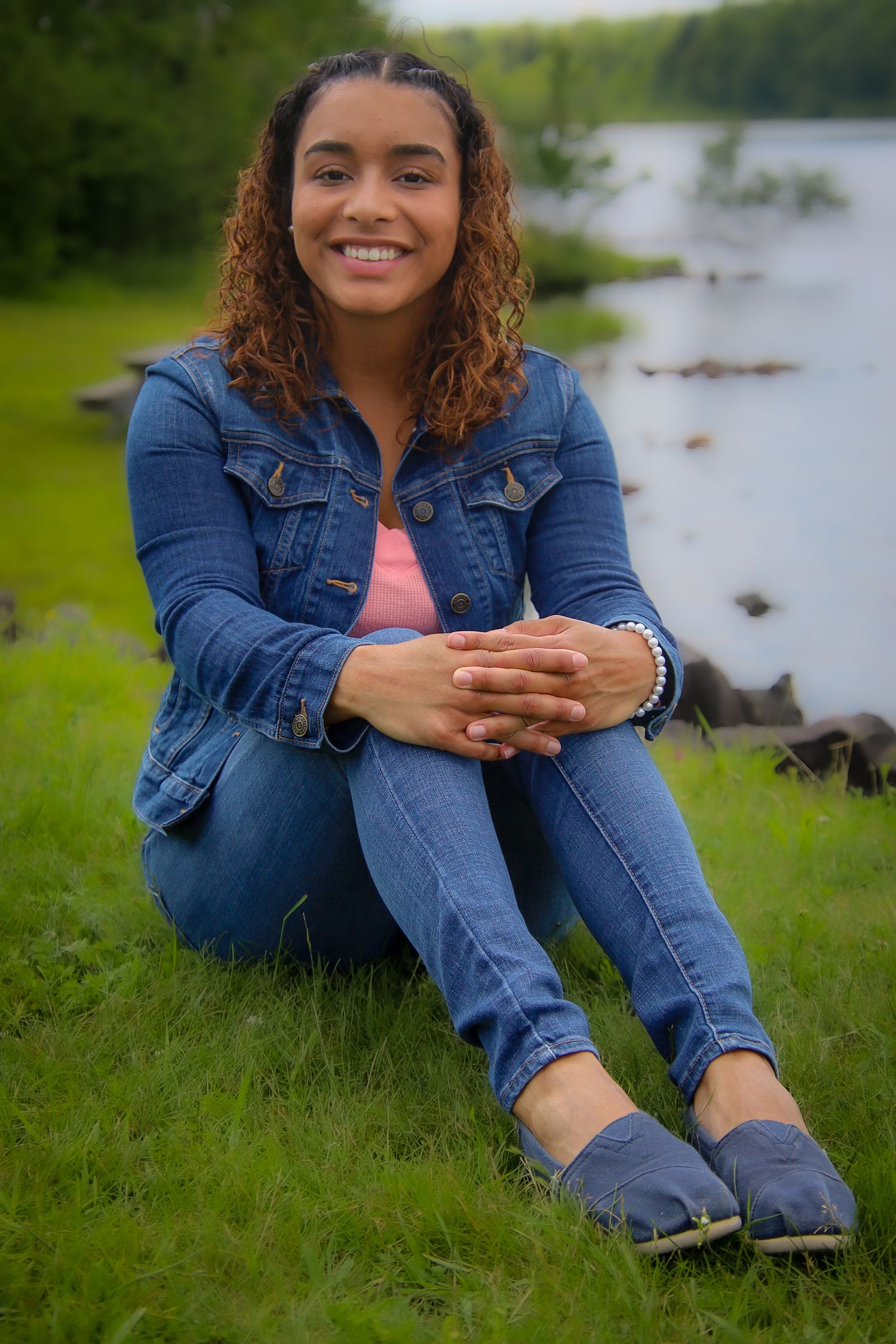 Person in denim jacket and jeans sits casually on grass near water in a series of outdoor portrait poses.