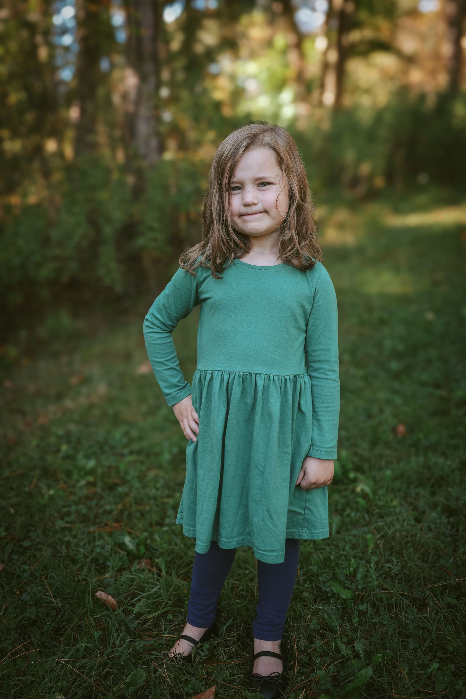 A young child wearing a green long-sleeved dress and navy leggings stands in a sunlit forest setting.