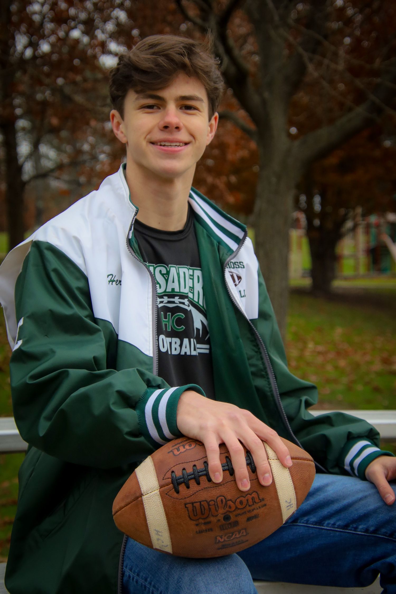 Student athlete in green and white varsity jacket poses with football on autumn day.