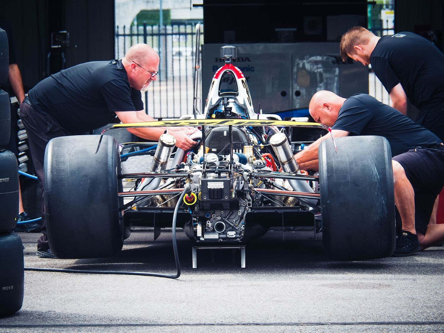Mechanics work on Formula 1 racing car in pit lane showing detailed view of rear wing and suspension components.