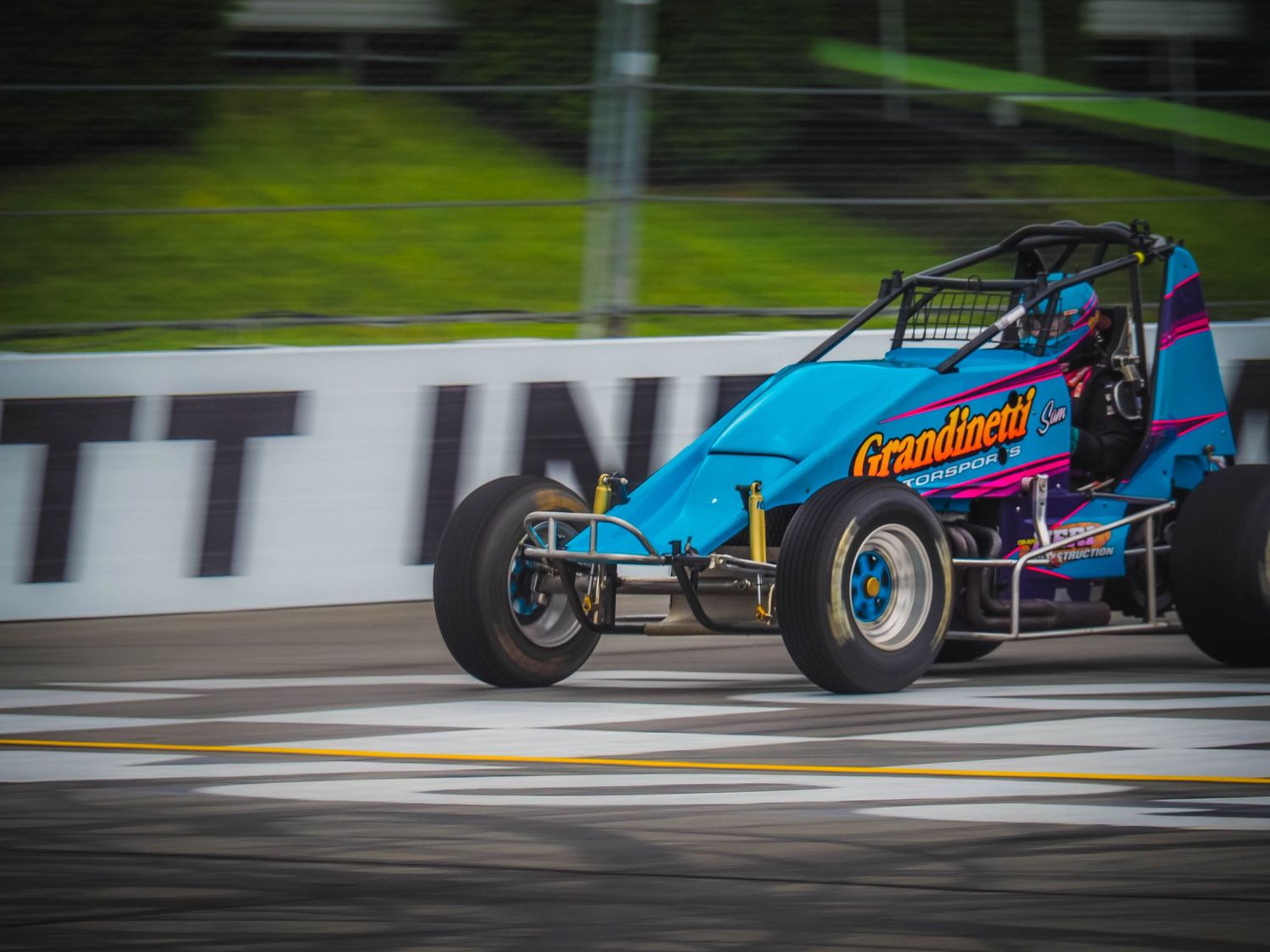A blue and pink sprint car races around a dirt track during a competitive motorsport event.