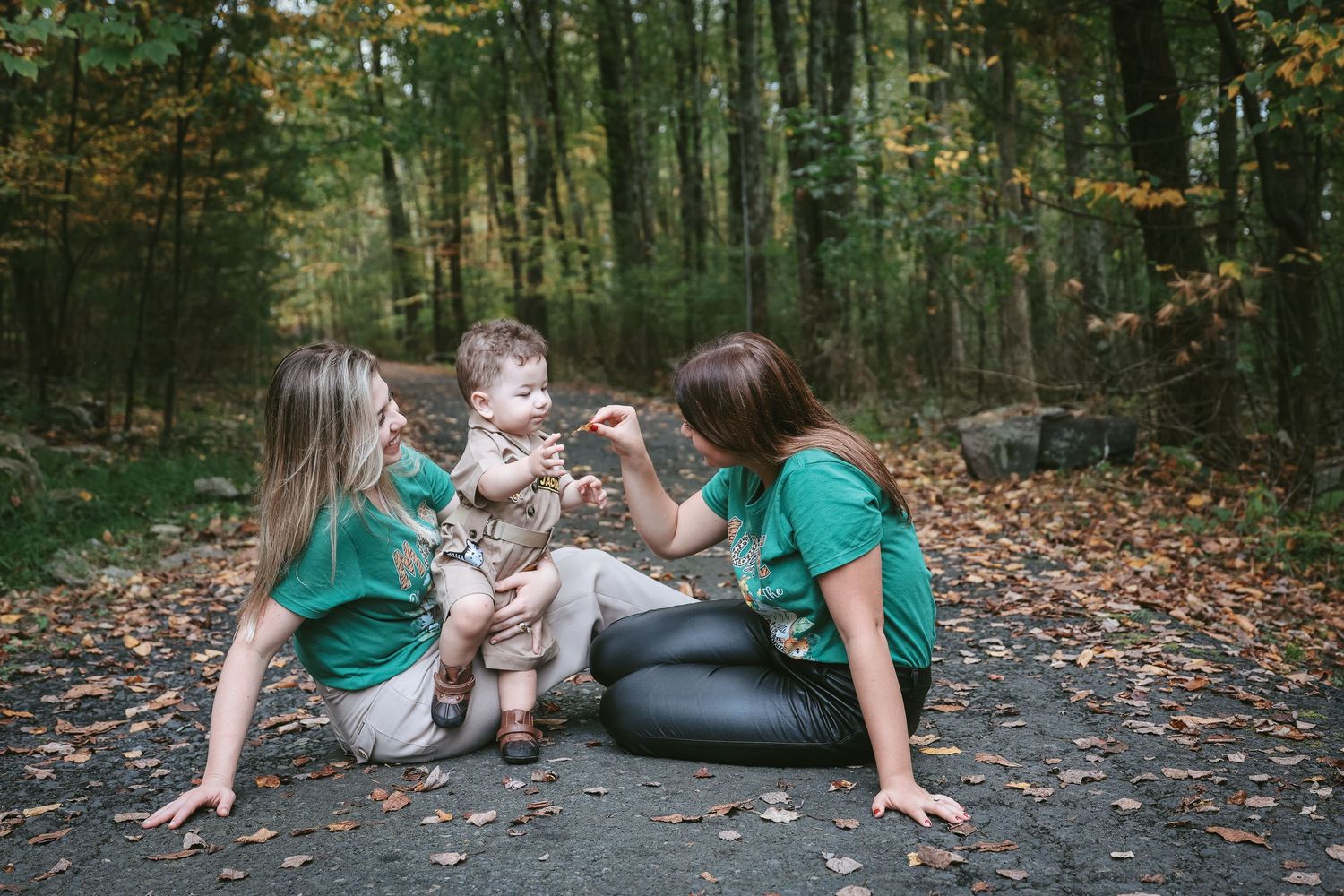 A family in matching teal outfits laying on autumn leaves in a wooded forest setting during a photo session.