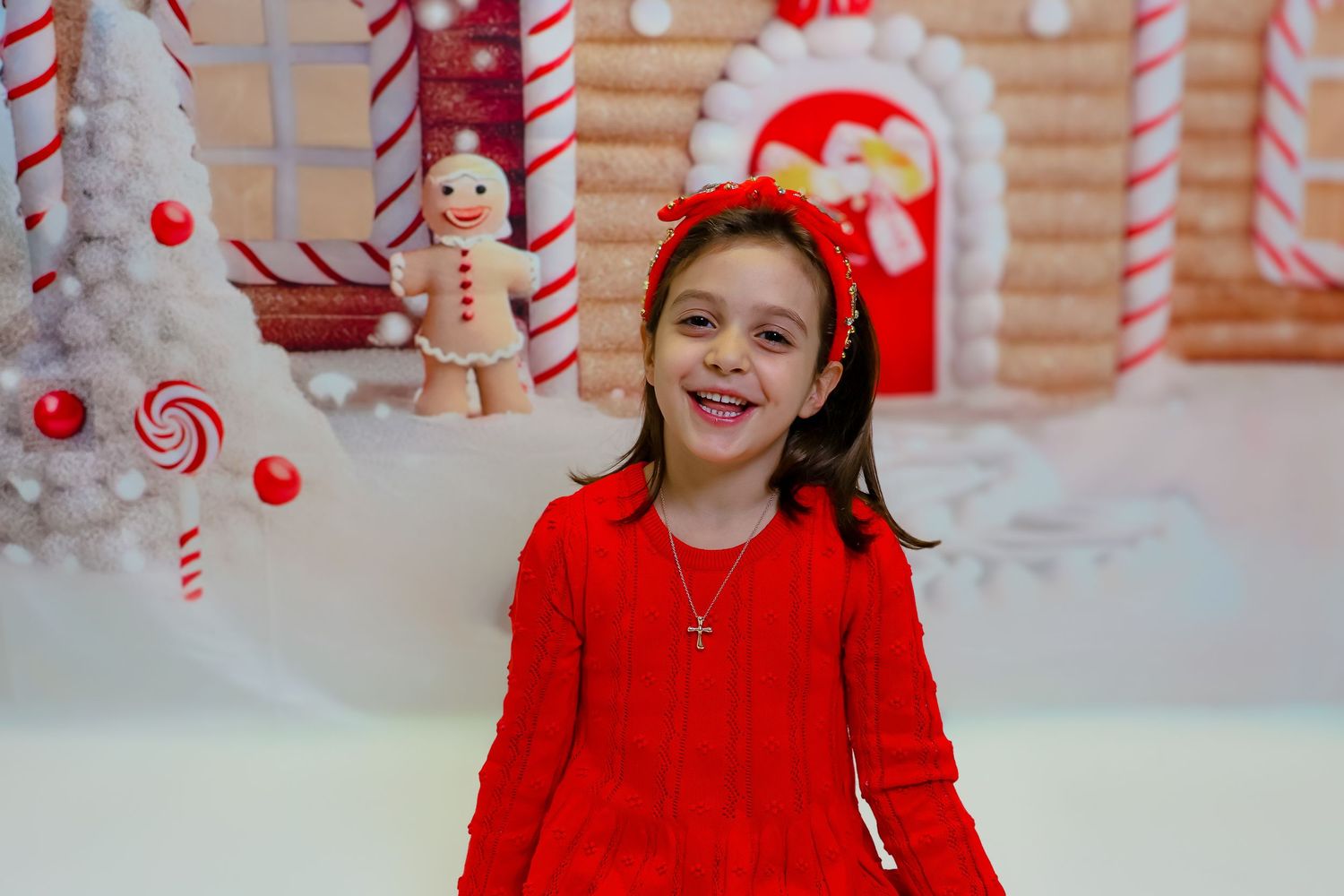 Child in red dress smiles and poses in front of a whimsical candy cane and snowflake holiday backdrop.