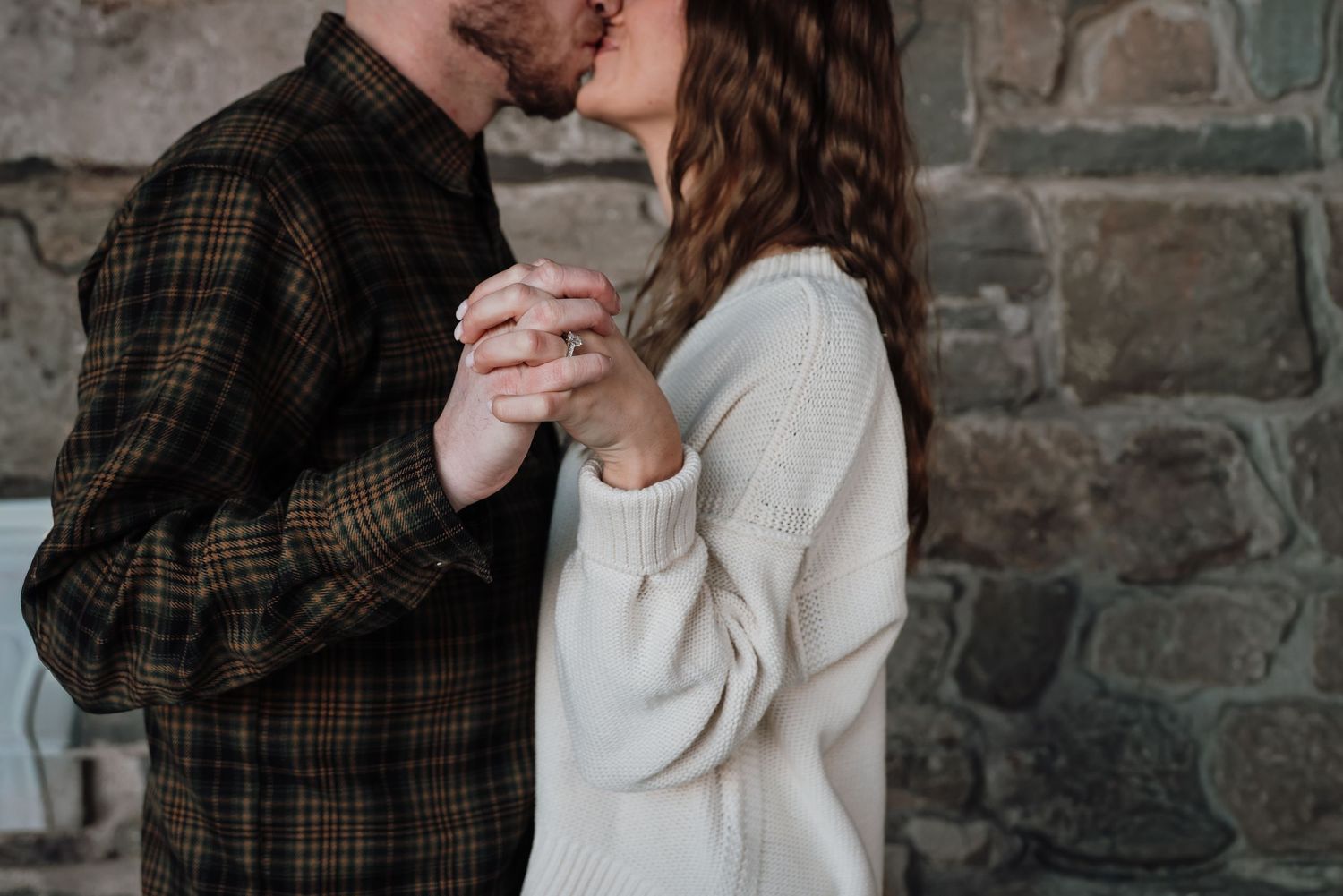 A couple holds hands intimately against a stone wall background, wearing casual sweaters in brown and cream colors.