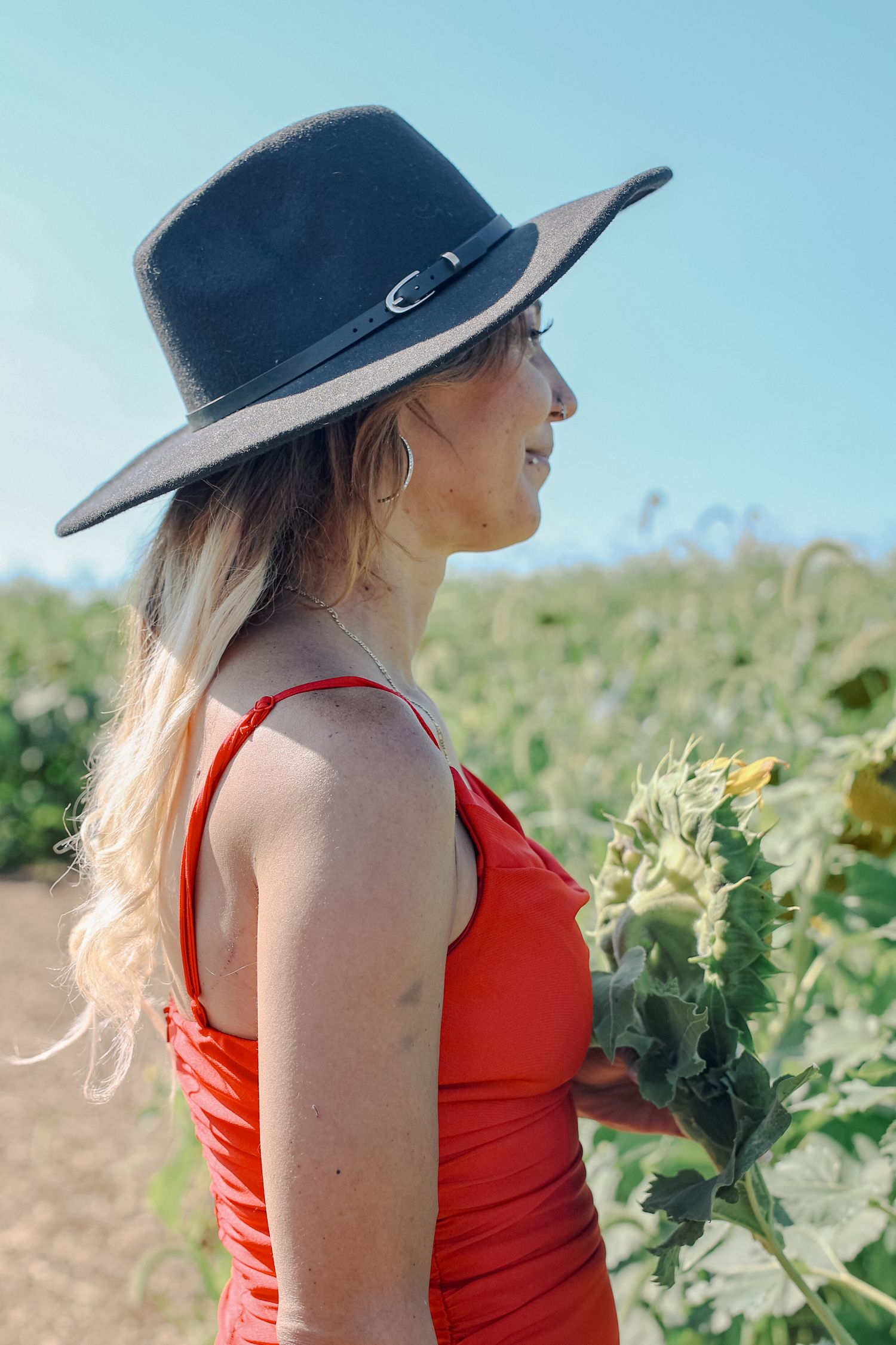 A person in a red tank top and wide-brimmed hat stands in a sunflower field on a sunny day.
