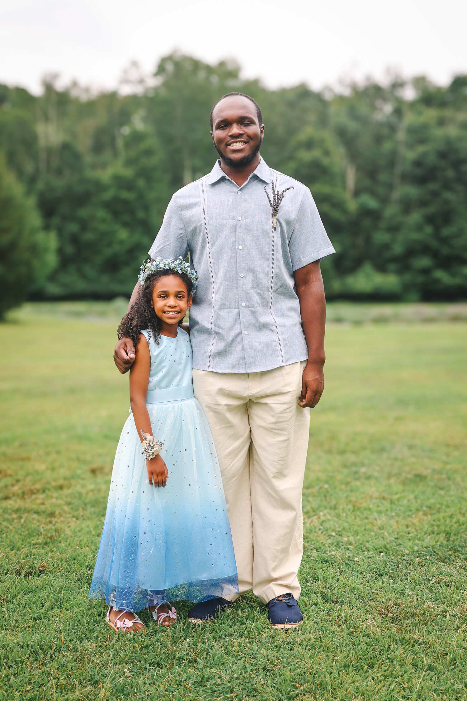 A parent and child in a blue tulle dress pose together on a grassy field during a summer photo session.