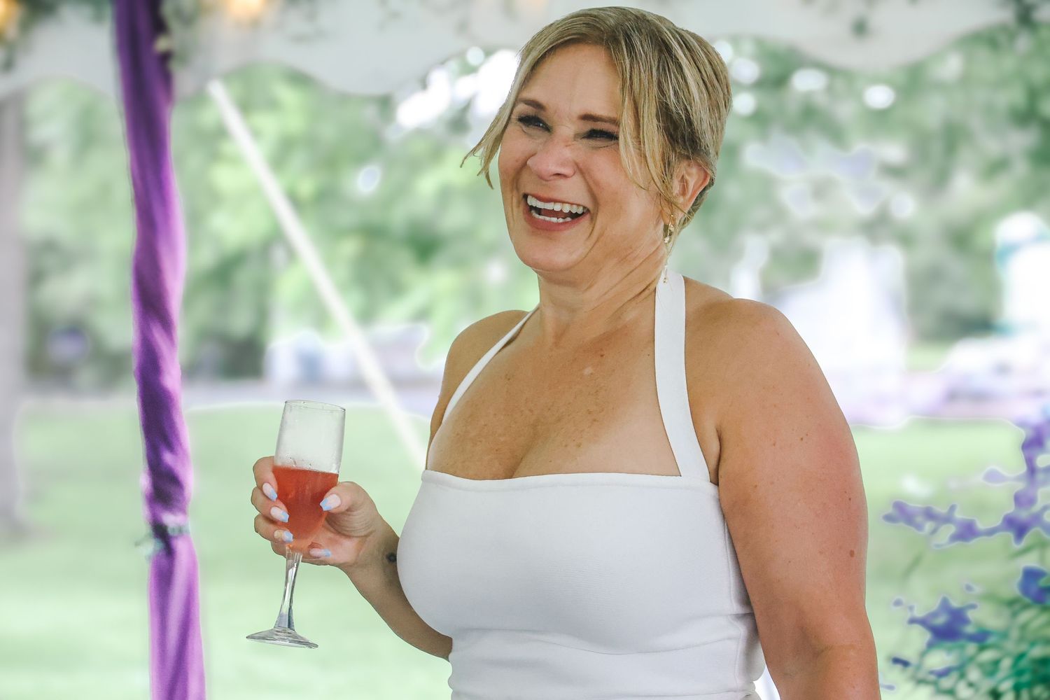 A person in a white dress holds a champagne glass and laughs during an outdoor celebration with purple decor.
