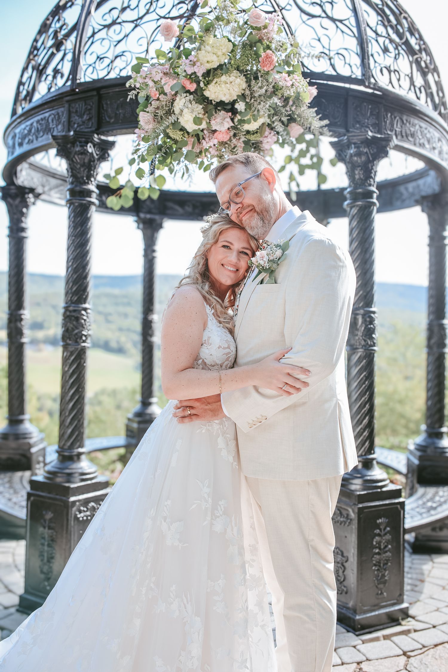 A romantic couple in wedding attire embraces under an ornate gazebo with mountain views in the background.