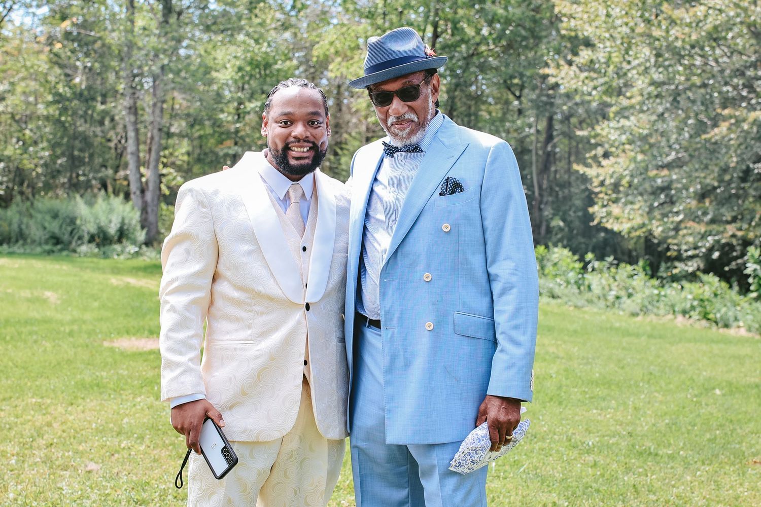 Two people stand together outdoors on a sunny day, one wearing a white suit and one in a light blue suit.