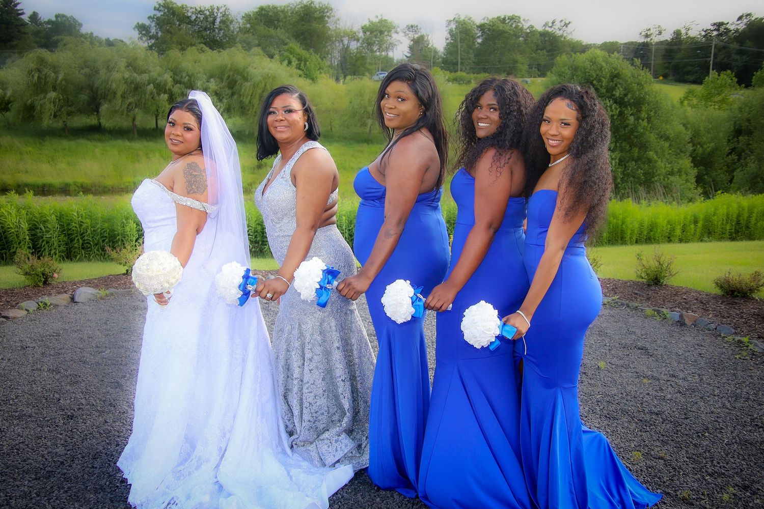 Wedding party in white and royal blue dresses pose together outdoors against a lush green landscape.