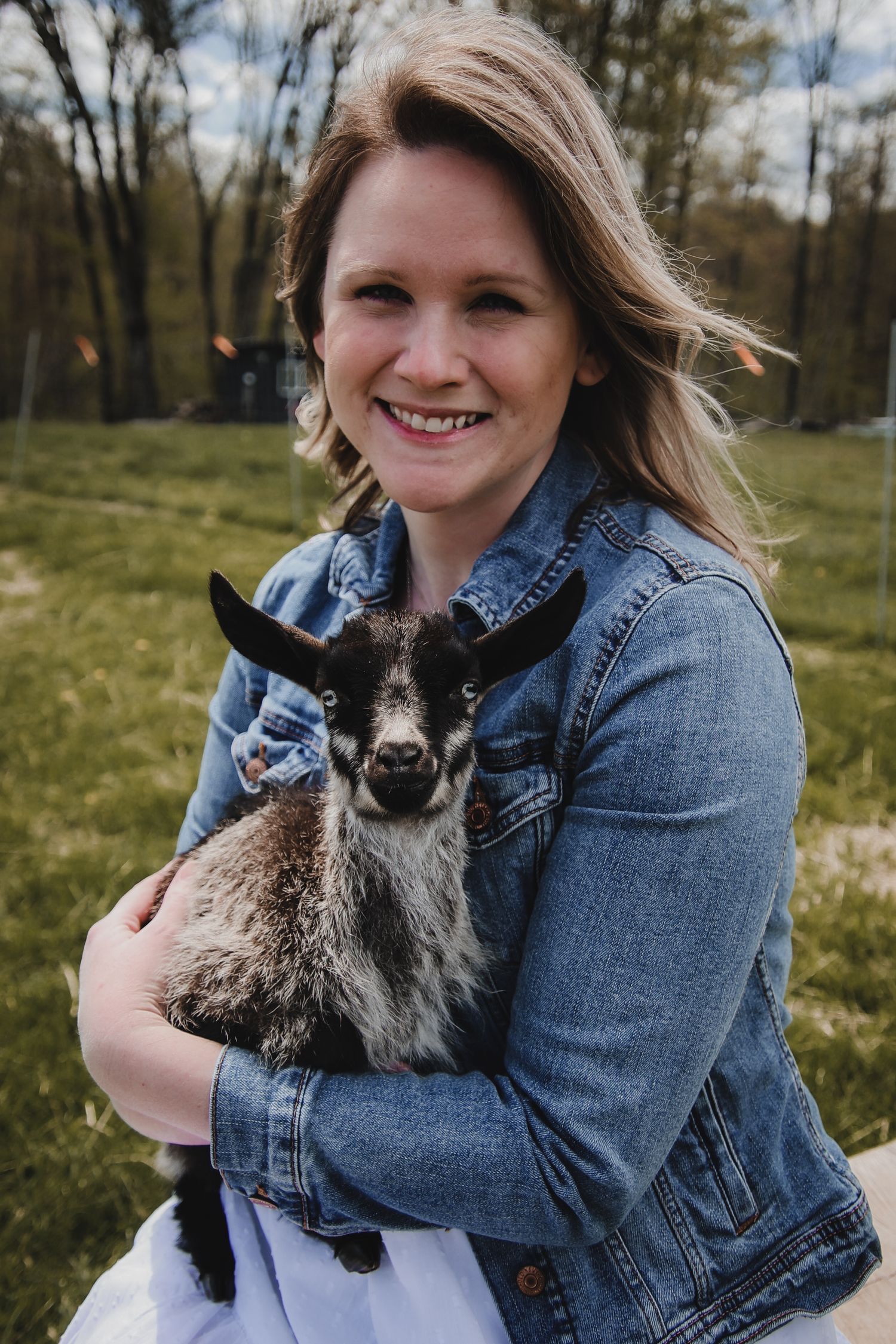 A person in a denim jacket holds a small fluffy black and white goat kid in a grassy outdoor setting.