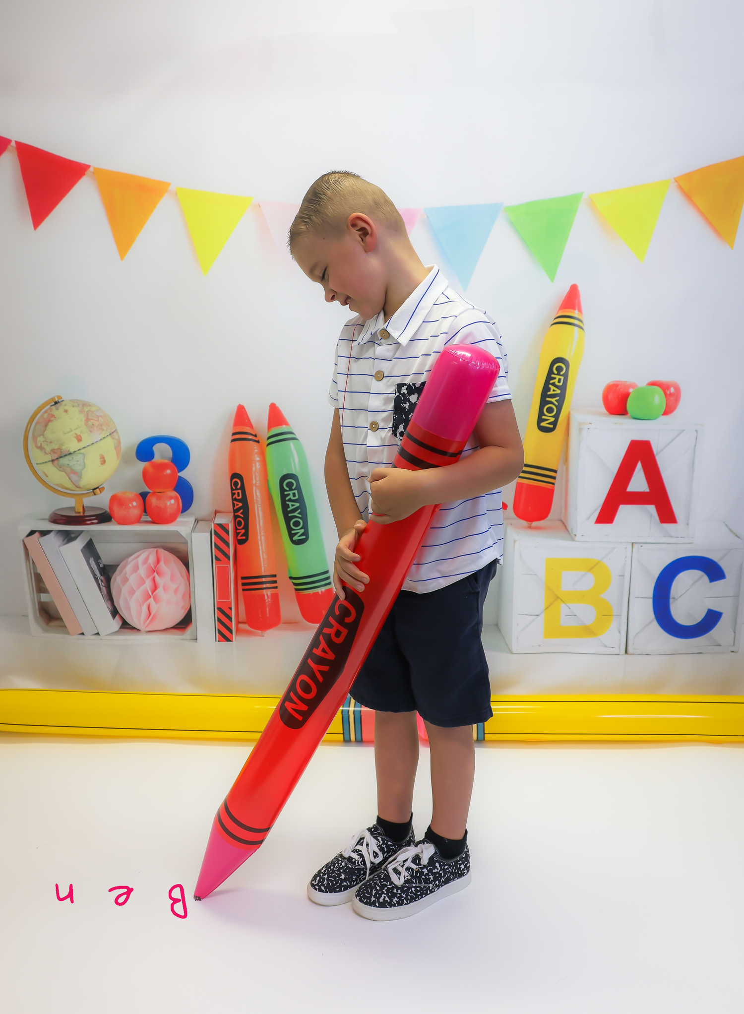 Young boy holding a giant red crayon prop in a colorful back-to-school themed photo with alphabet blocks and bunting.