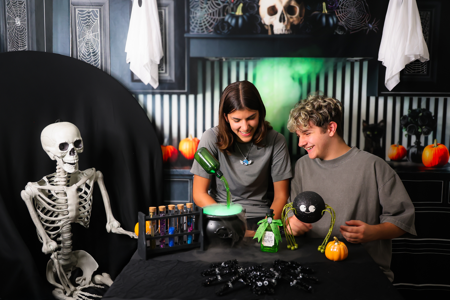 Two teens craft Halloween decorations at a spooky table with skeletons, test tubes, and black balloons.