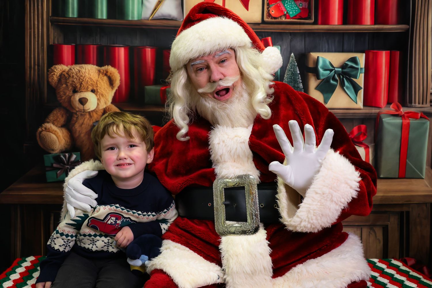 Young child sits with Santa Claus in festive holiday setting with teddy bear and wrapped gifts in background.