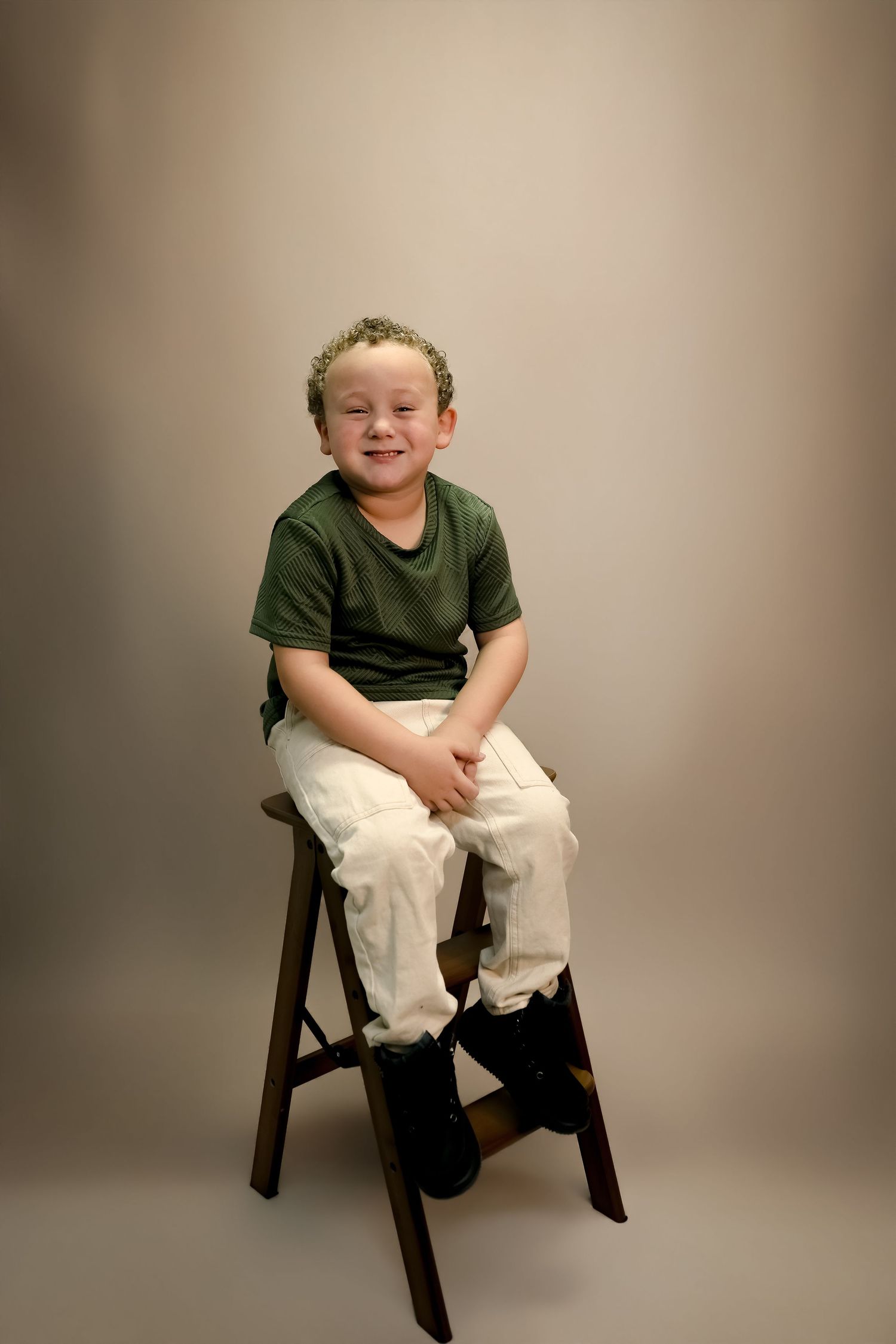 A smiling young boy with curly hair sits on a wooden stool wearing a green t-shirt and white pants in a studio portrait.