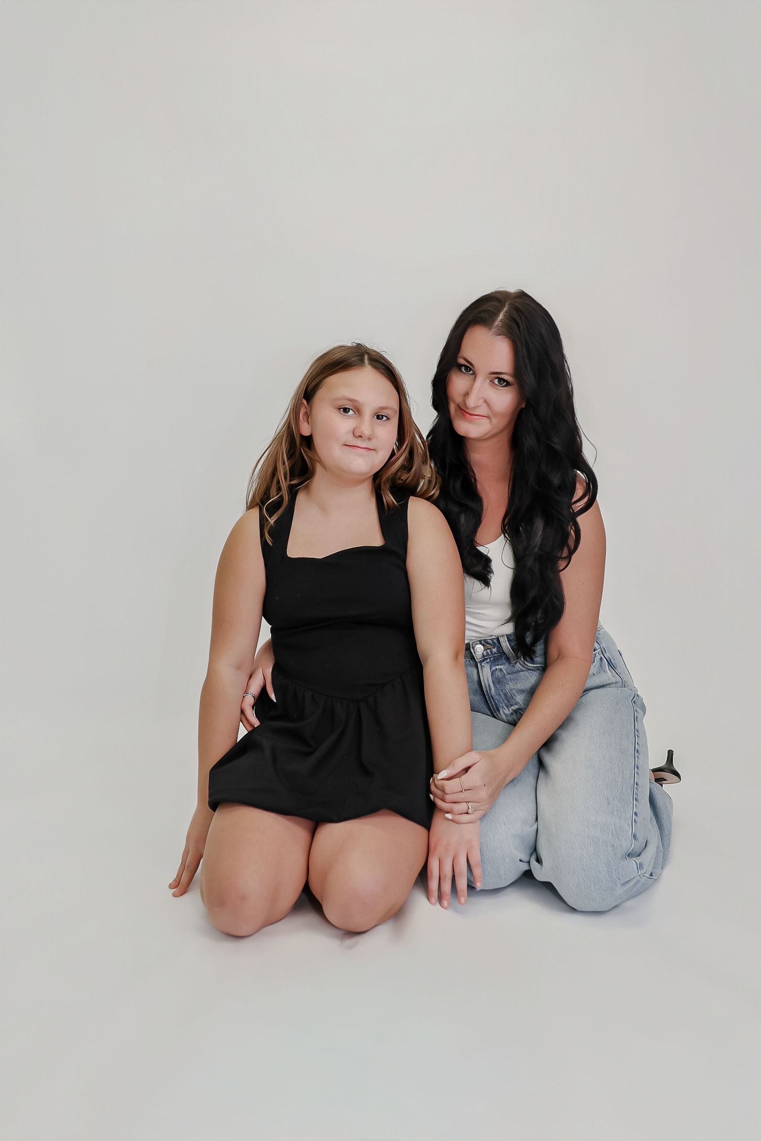 Two girls sitting on floor, one in black dress, one in white top and jeans, posing for a studio portrait.