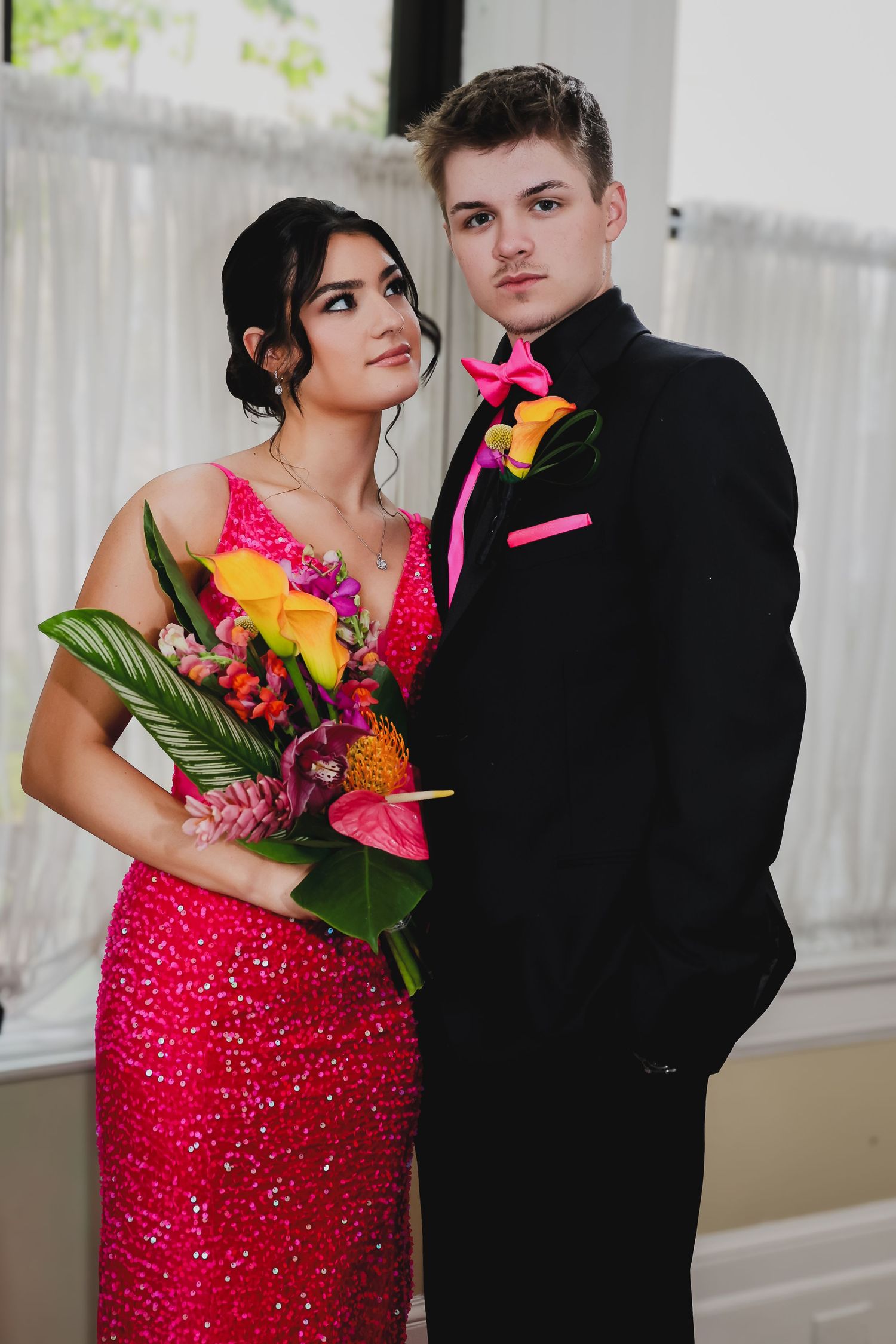 A couple dressed for prom, woman in pink sequin gown holding tropical bouquet, man in black tuxedo with pink boutonniere.