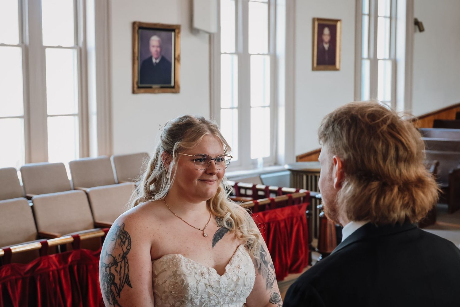Bride in lace gown and glasses exchanges vows with partner in elegant wood-paneled ceremony hall with portrait paintings.