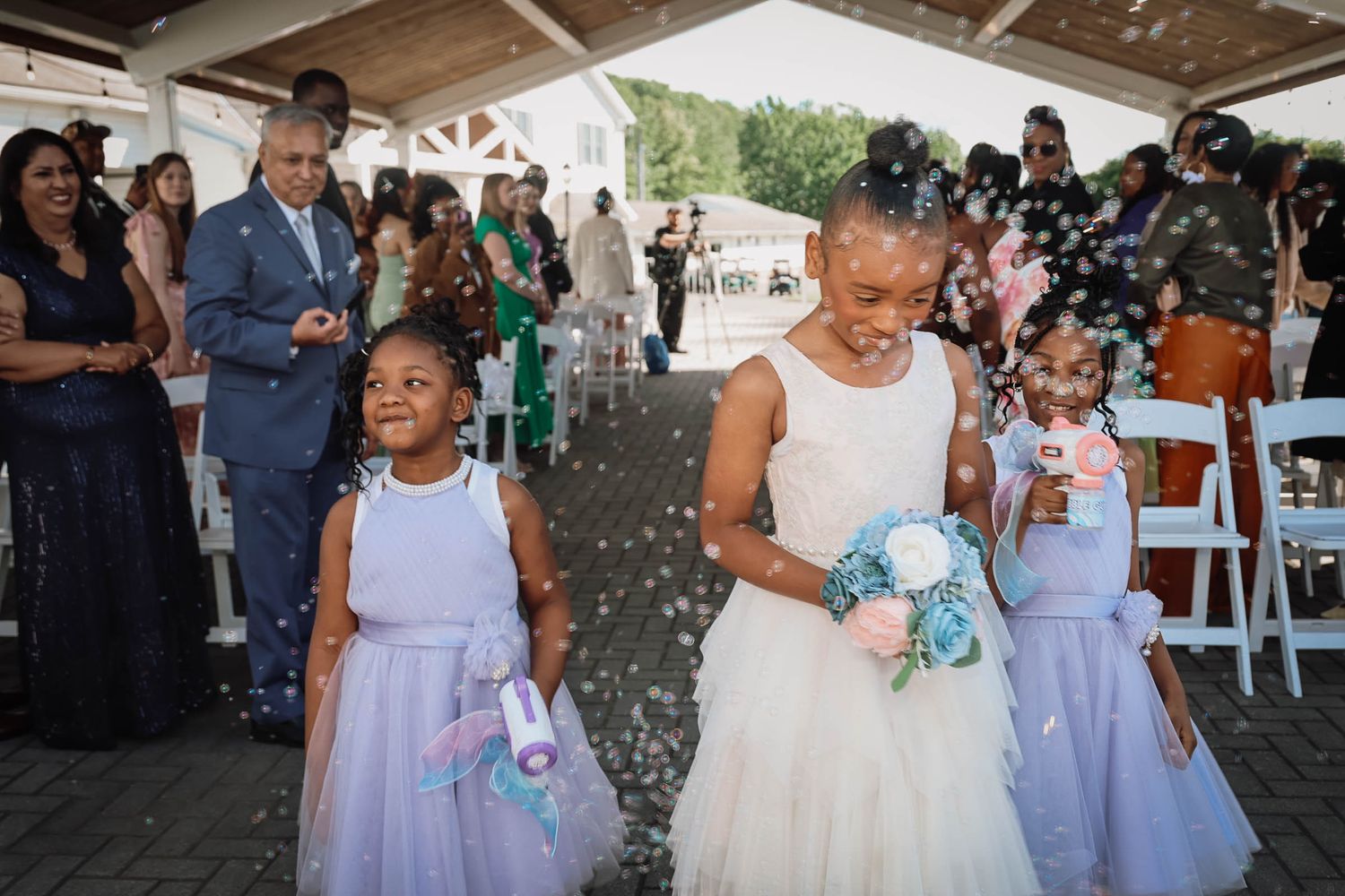 Bride walks with flower girls through bubble shower at outdoor wedding ceremony, guests applauding in background.