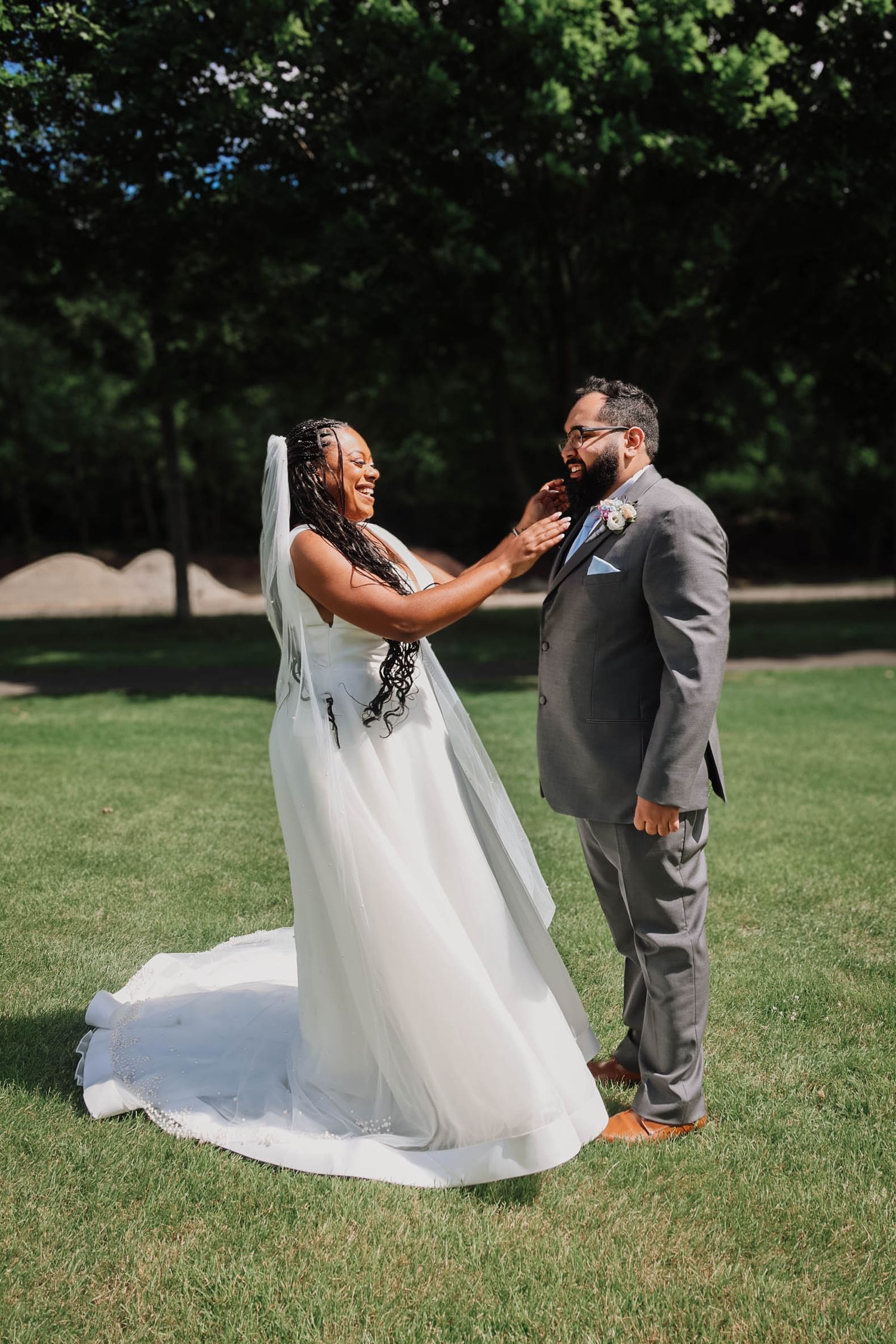 Bride in white gown and veil touches groom's face as they share a tender first look moment on a sunny green lawn.