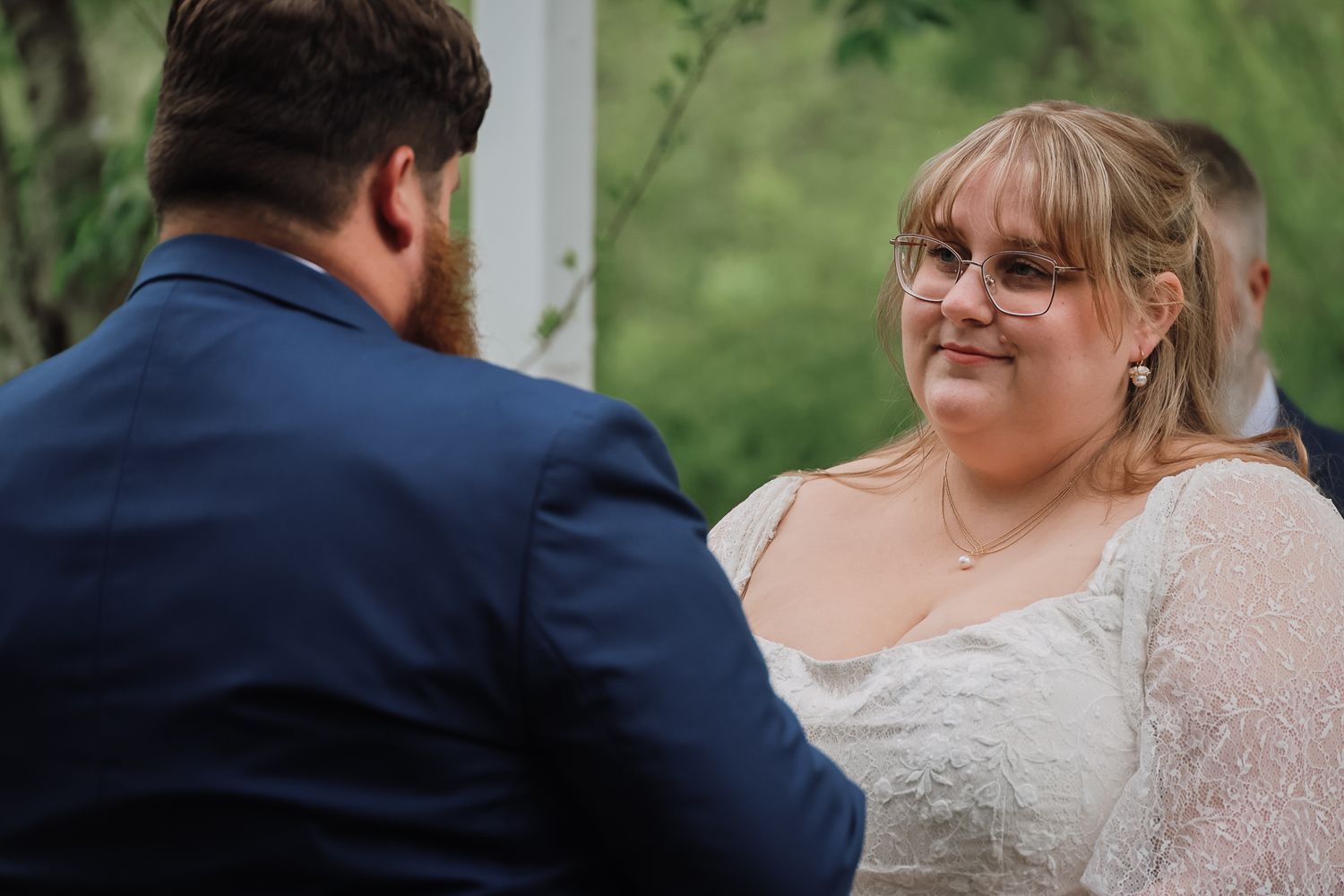 Bride in lace wedding dress smiling at groom in navy suit during outdoor wedding ceremony.