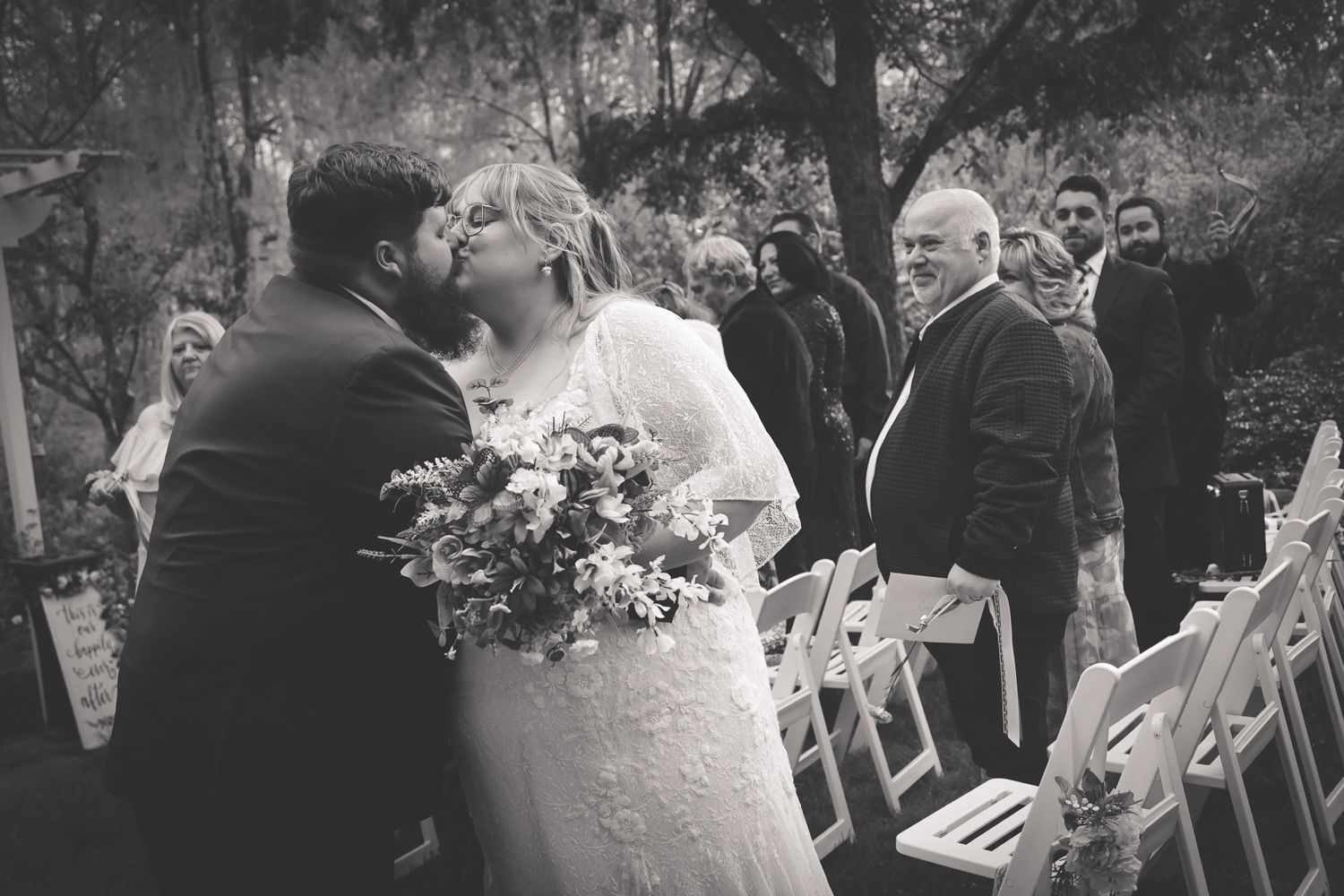 Black and white wedding photo of bride and groom sharing a kiss at outdoor ceremony aisle with guests watching.