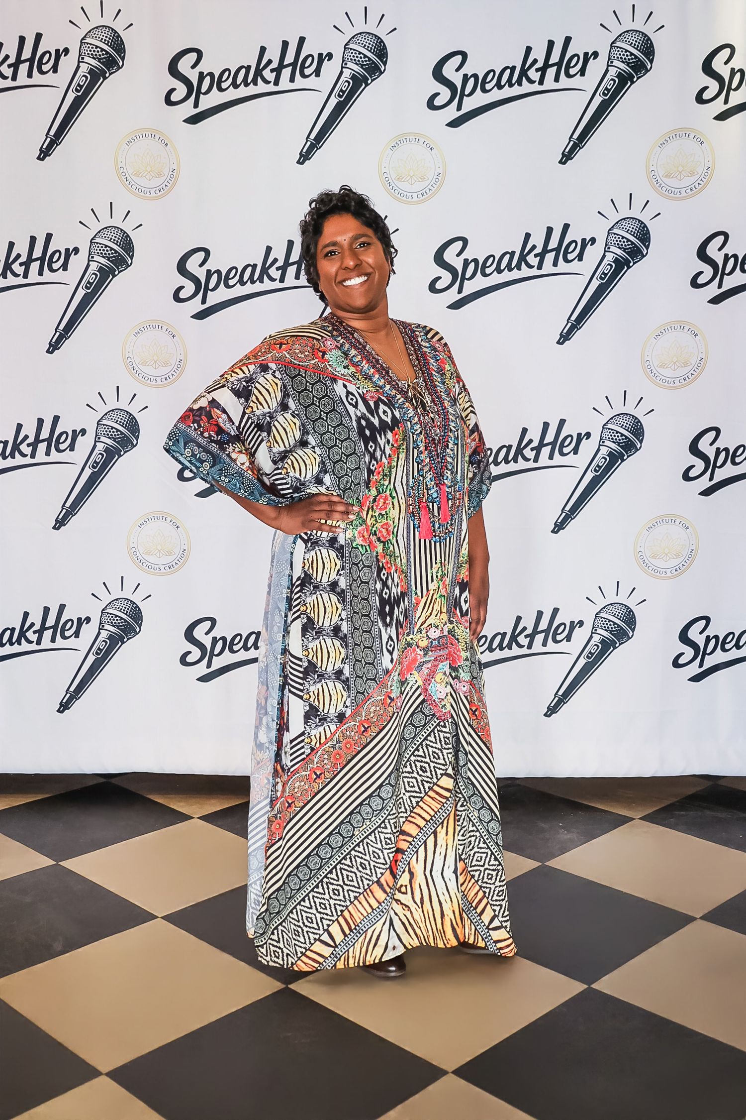 Woman in colorful patterned maxi dress poses at SpeakHer event backdrop with microphone logos.