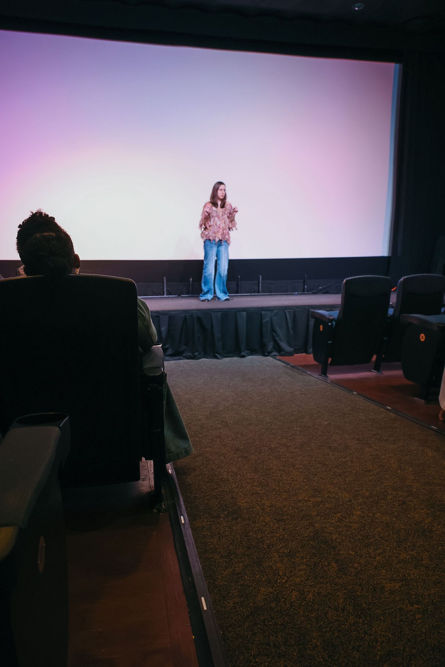 A woman stands on a dark stage before a large screen, addressing a small audience in a theater.