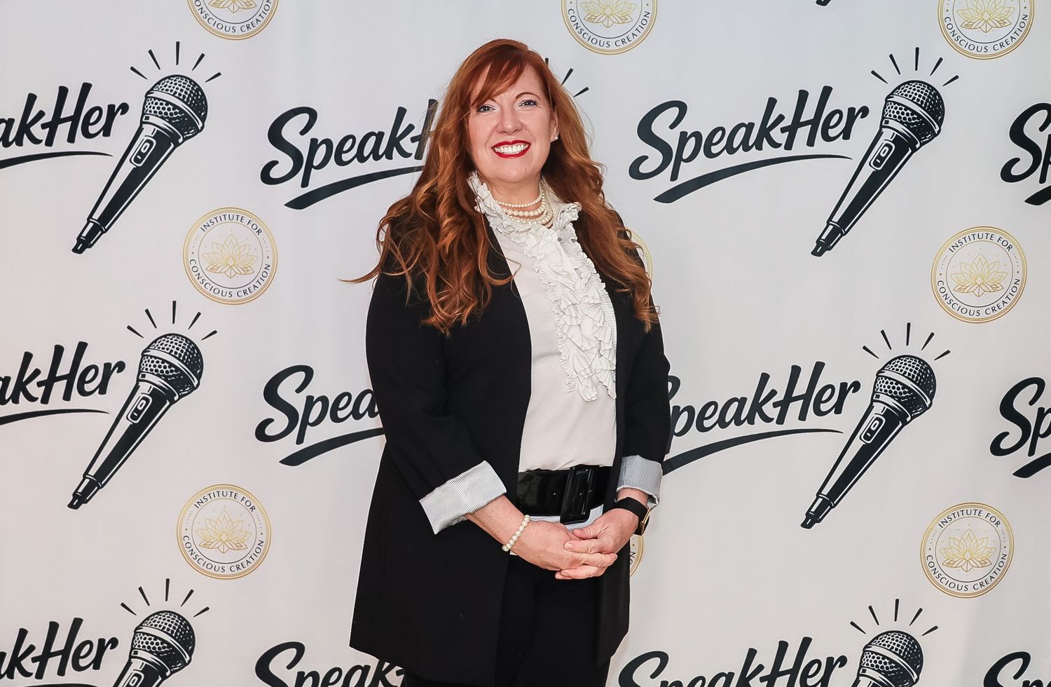 Woman in black blazer posing at SpeakHer event backdrop with microphone logo branding.