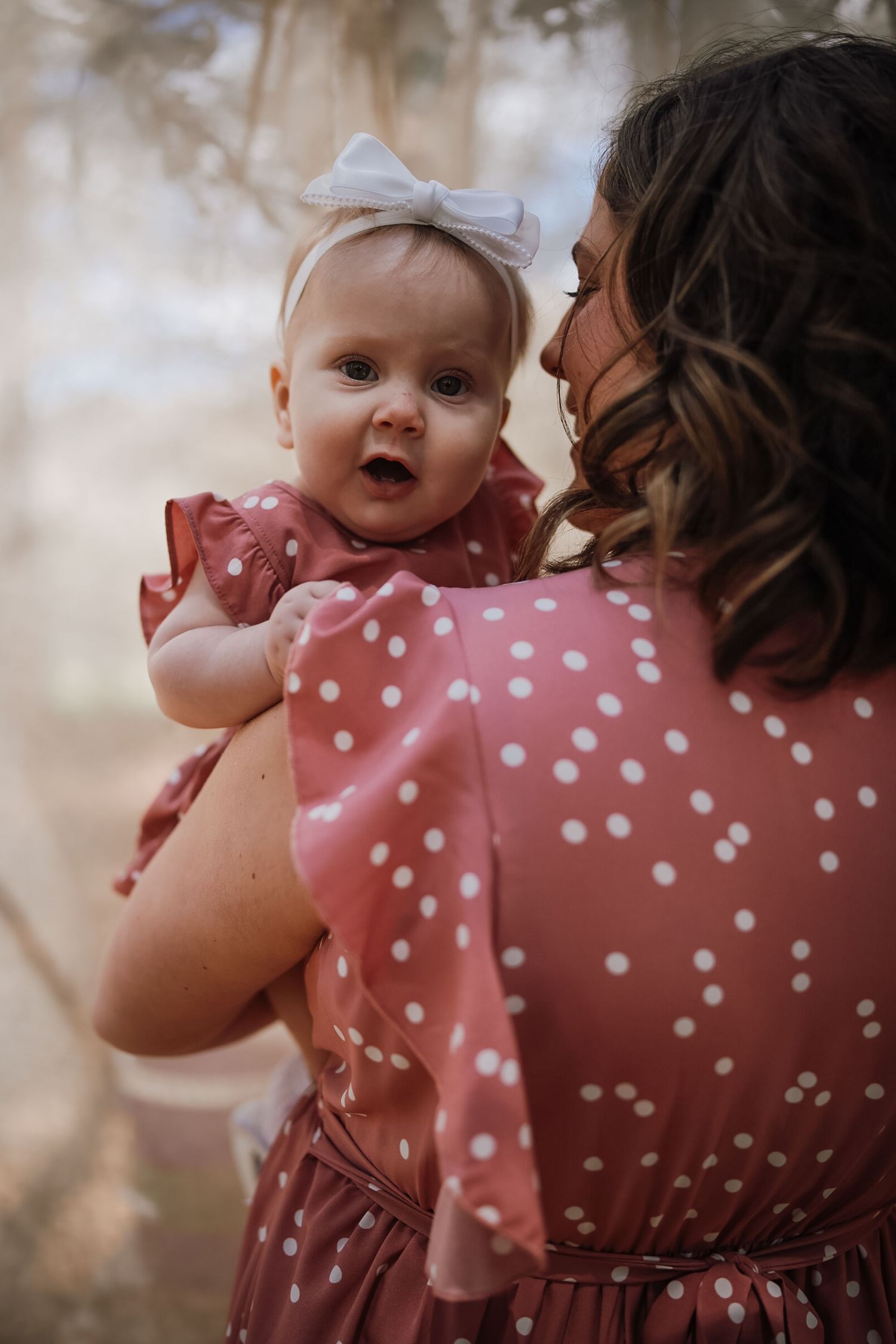 A mother in a polka dot dress holds her baby girl wearing a matching dress and white bow headband outdoors.