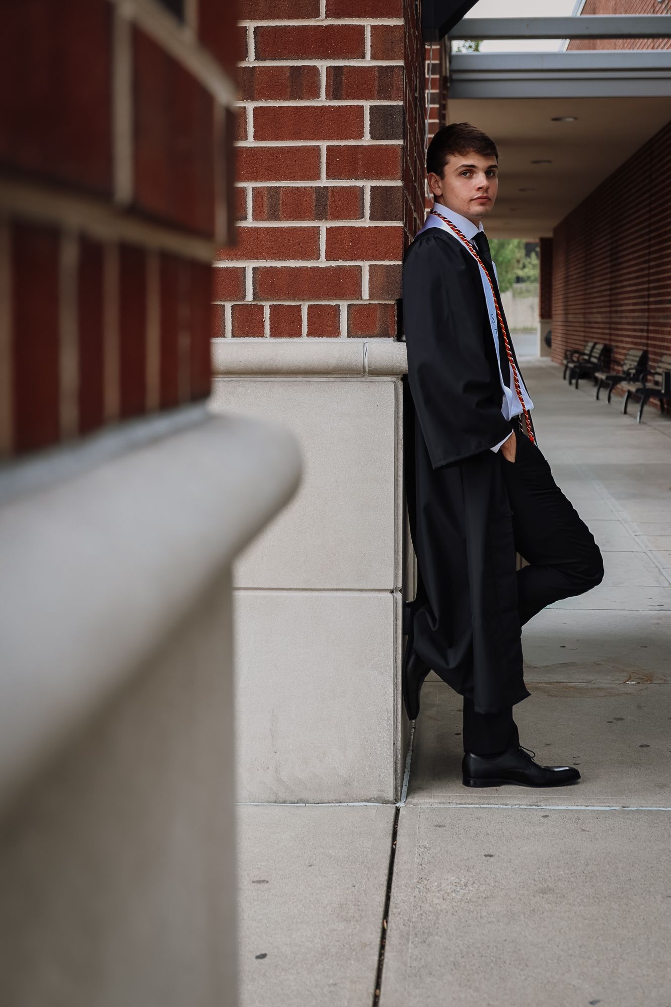 Young man in graduation gown and stole leaning against brick wall outdoors on campus sidewalk.