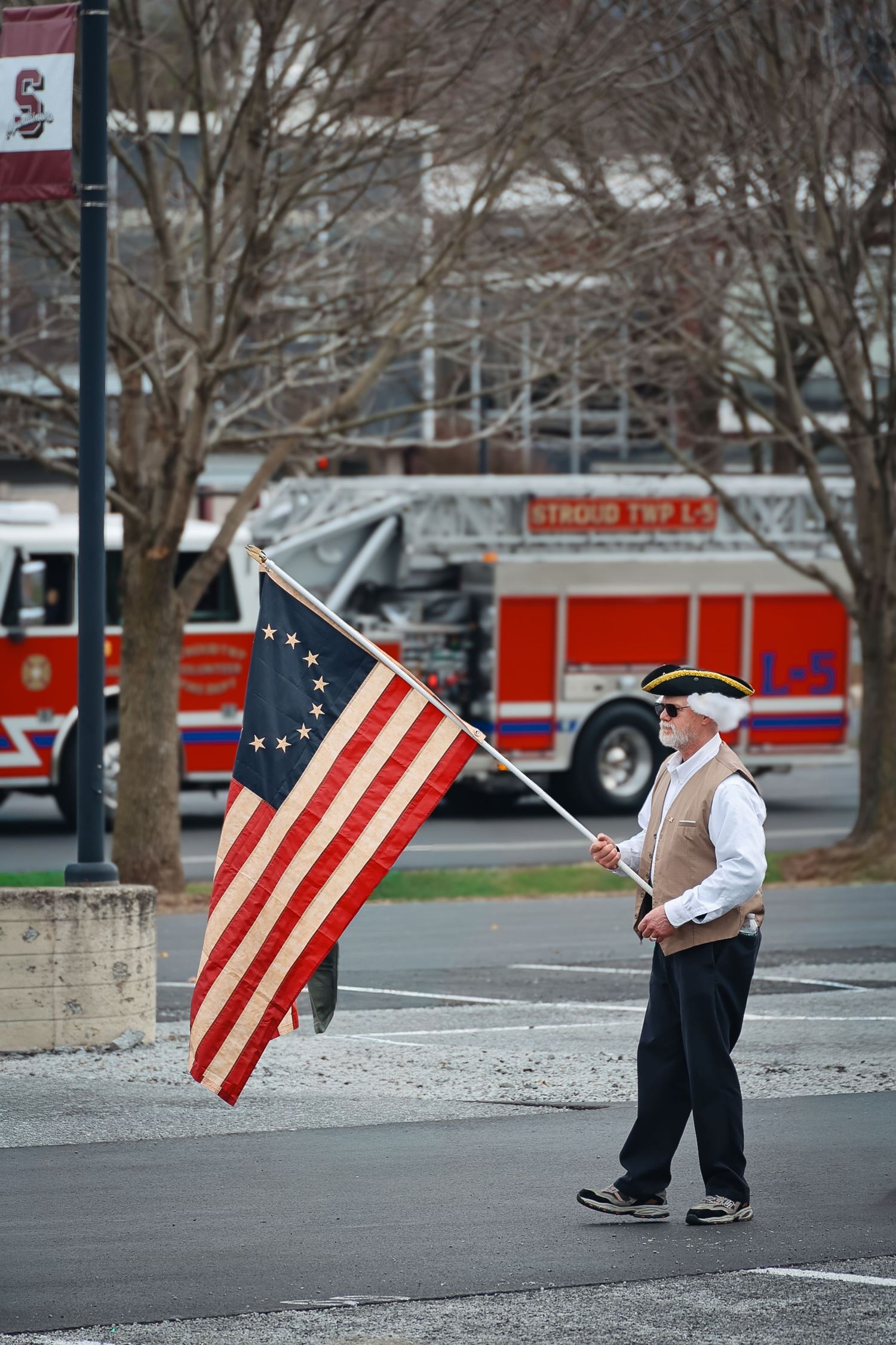 Man in colonial costume carrying a Betsy Ross flag during a parade, with a fire truck visible in the background.